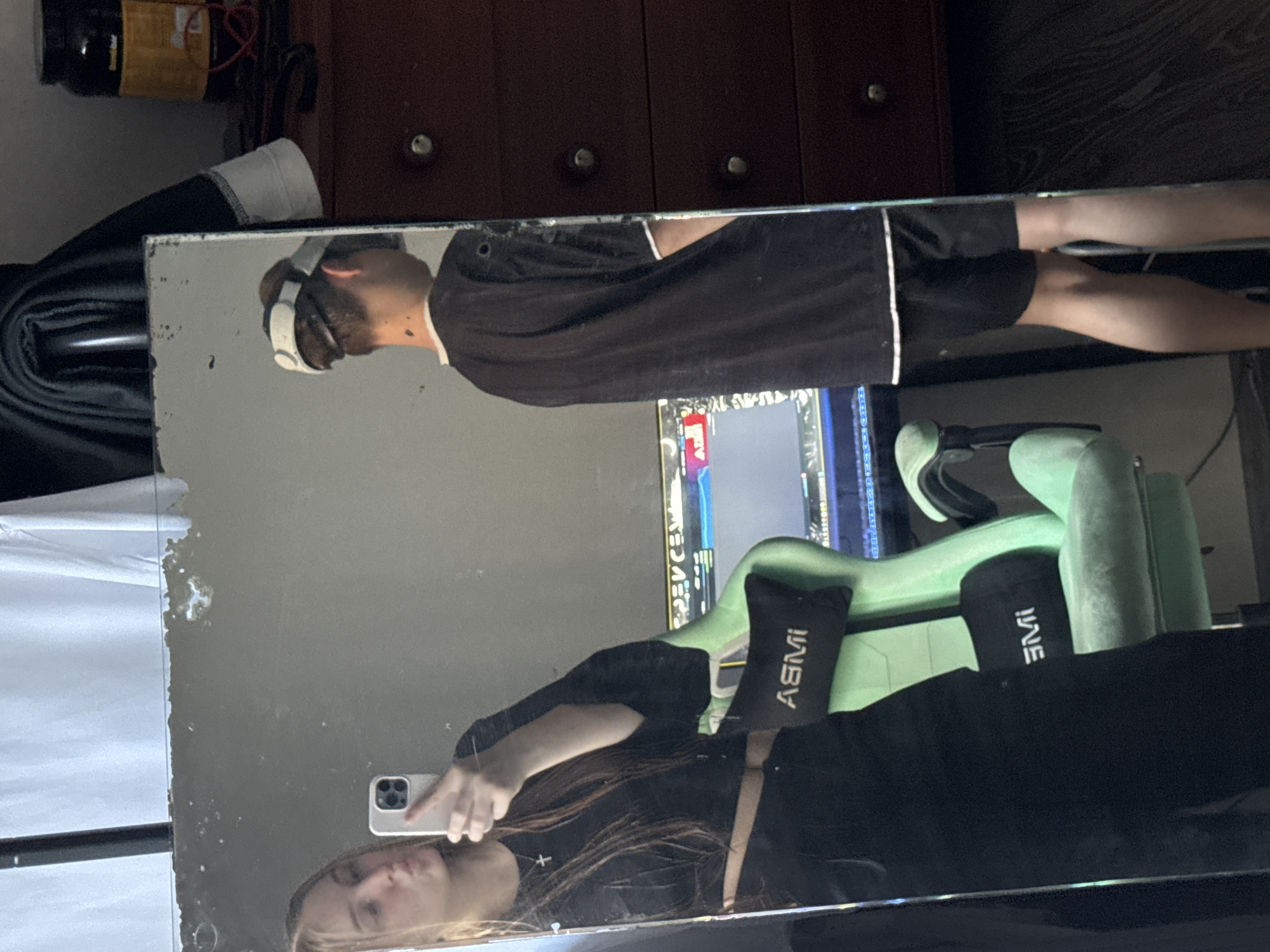 An indoor, vertically oriented mirror selfie captures two individuals and their surroundings in Perm, Russia. The image is taken by a person, likely female, holding a white smartphone, whose reflection is visible in the lower left portion of a large, aged mirror. This individual has long, light brown hair and wears a dark top, with a faint cross visible on a necklace.
Reflected in the upper part of the mirror is another person, likely male, wearing white headphones, a black t-shirt, and black shorts with white trim. This person stands in front of a light green gaming chair, which features black lumbar and headrest pillows. The lumbar pillow has "ASMI" printed in white text, while the headrest pillow shows "IME". Behind the standing person, a computer monitor displaying a digital interface is visible.
The background above the mirror includes a dark wooden dresser or cabinet with silver knobs, and a black container with a yellow label on top. The mirror itself exhibits some deterioration along its left edge, indicating age or wear. The overall scene suggests a casual home environment, possibly a bedroom or gaming setup. The lighting is artificial, giving no clues about the time of day or outdoor weather.