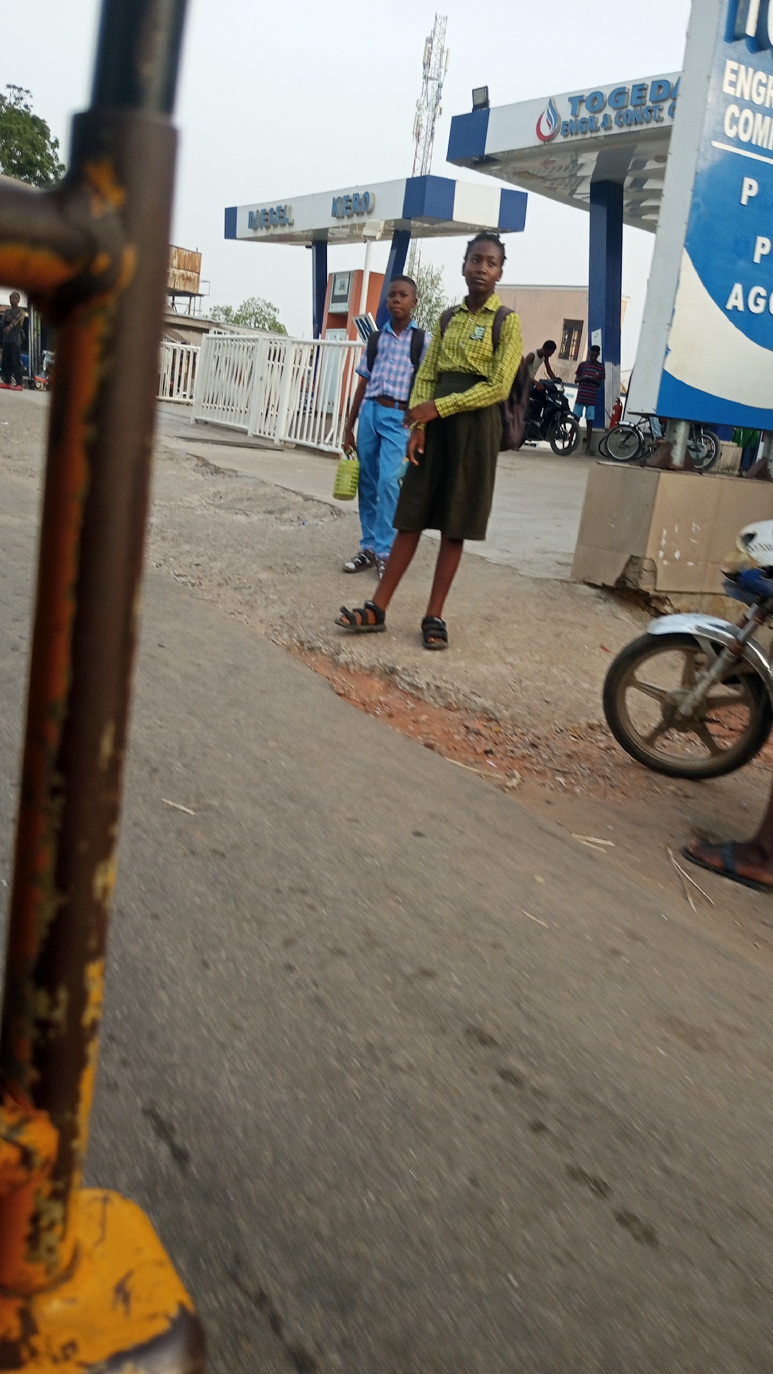 The image, captured in Zango-ndaji, Nigeria, shows two school children standing on a roadside near a Togeida energy company gas station. The girl in the foreground wears a yellow shirt and dark skirt, with a backpack. The boy behind her wears a blue and white checkered shirt, blue pants, and carries a green container. Both wear sandals. To the side, there is a motorcycle and bicycles. The foreground includes part of a yellow object with rusted metal. The road surface is asphalt with debris. In the background, there are other individuals, a building, and communication towers.