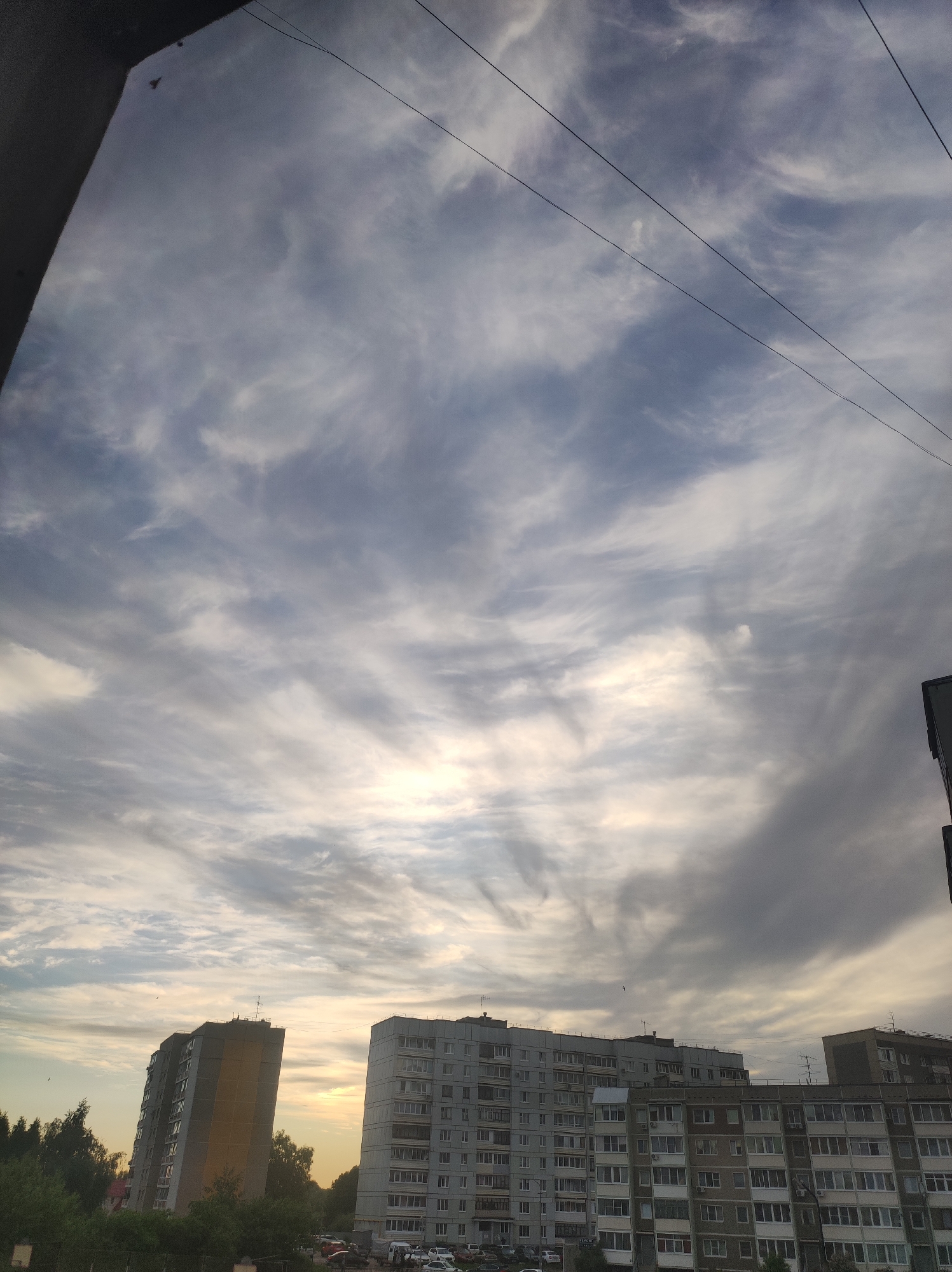 The image shows a view of a neighborhood in Chaikovsky, Russia, at either sunrise or sunset. In the foreground, multiple Soviet-era apartment buildings are visible. Cars are parked in the spaces in front of the buildings. Behind the buildings are trees with green leaves. Above, the sky is filled with altocumulus clouds, appearing striped, with a golden glow from the sun near the horizon. Three power lines are strung across the upper portion of the sky. The edge of a building is visible on the left and right edges of the frame.