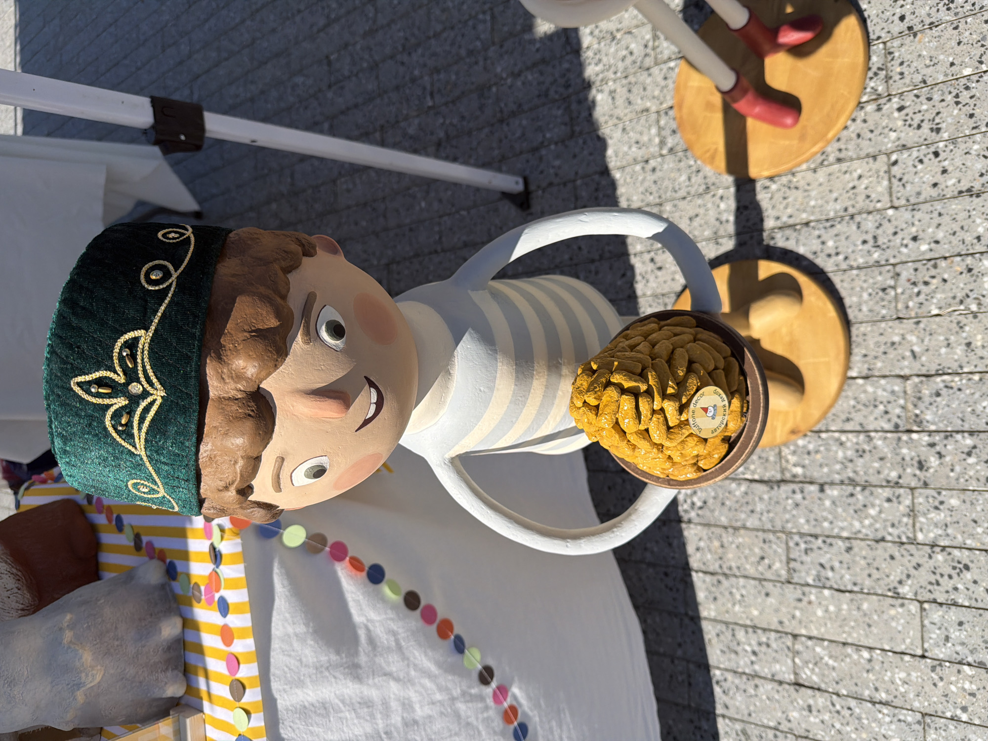 This is a full-body, eye-level, outdoor, medium shot of a doll-like figure standing on a paved surface. The figure is a child with brown hair, light skin, and large, round eyes. It wears a green, felt hat with gold embroidery and a white and light blue striped shirt. The figure holds a round, brown bowl filled with yellow, honeycomb-shaped objects, which appears to be a honey cake. The doll stands on a circular wooden base, and its legs extend down to a second, larger circular base. To the right of the figure, another doll's legs, clad in red boots, are visible, also standing on a circular wooden base. In the background, a white tent or canopy pole is visible, along with a table covered with a white cloth and a yellow and white striped runner. A garland of colorful paper circles hangs from the table. The ground is paved with light-colored square tiles. This scene is located in Kazan, Russia, likely at an outdoor market or festival, given the presence of vendor tents and decorated displays.