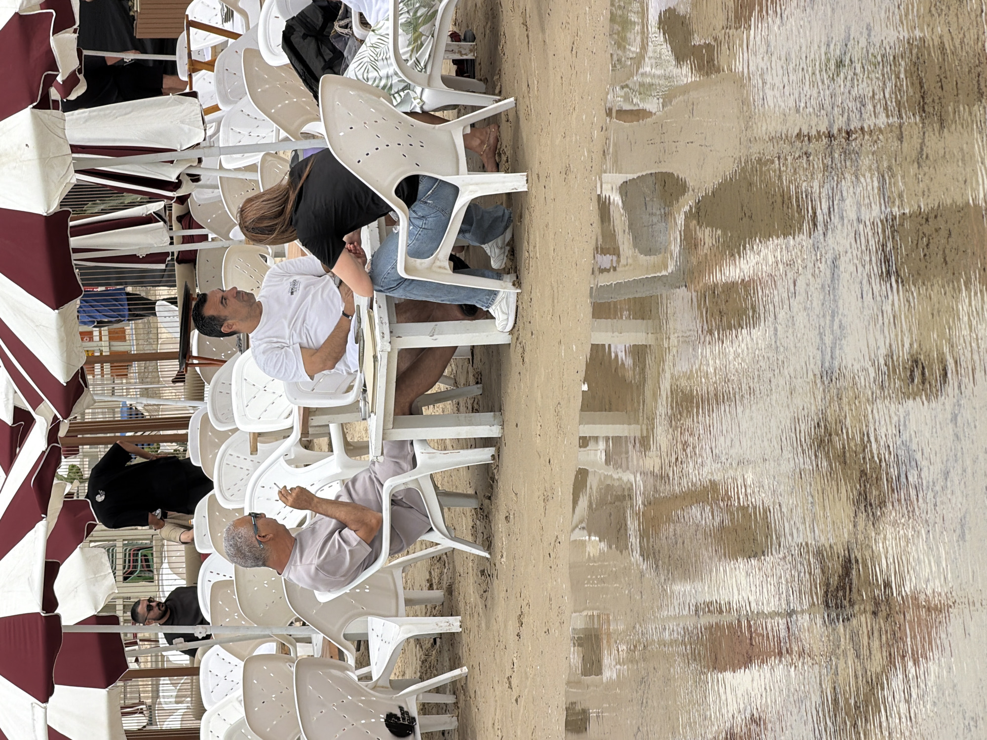 The image depicts an outdoor seating area, possibly a beachside cafe or restaurant, located in Tel-Aviv, Israel. Several white plastic chairs are arranged around tables under striped umbrellas. People are seated at the tables. One man wears a white t-shirt and shorts, while a woman in a black t-shirt stands beside him. Another man, wearing sunglasses and a light-colored shirt, sits at a nearby table and is holding a cigarette. A man wearing sunglasses can be seen sitting towards the lower left. The background shows a beach or shoreline with water that creates a reflection.