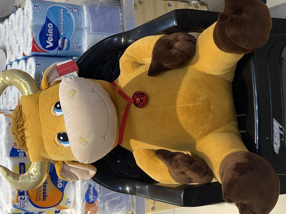 A large plush toy, resembling a golden-yellow bull or cow with light beige snout, blue eyes, and golden horns, is seated upright in a black plastic chair. The toy wears a red ribbon around its neck, from which a glossy red spherical bell hangs. Its hooves, on both hands and feet, are dark brown. A red-and-white tag, partially visible on its left ear, displays text including "2029 * 00". In the background, multiple large bundles of toilet paper are stacked, forming a retail display. Visible branding on the packaging includes "Veiro ДОМАШНЯЯ" (Veiro DOMASHNYAYA) with a blue label and rabbit graphic, and "Veiro classic" on a white label, with Cyrillic text visible on both. The scene is located within a retail establishment in the city of Golikovo, Russia.