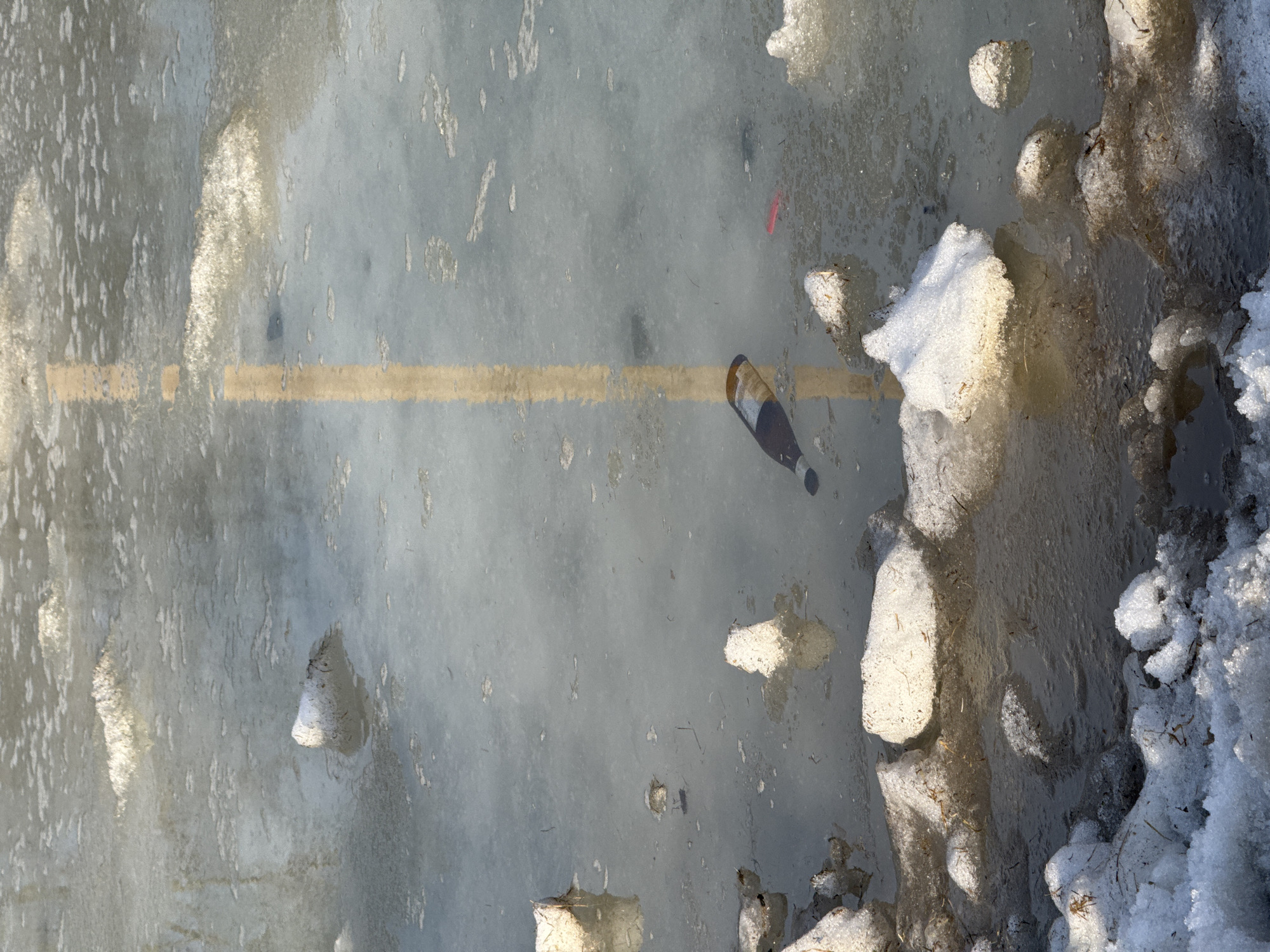 The image shows a patch of melting snow and ice, possibly on a street or sidewalk in Izhevsk, Russia. A discarded brown glass bottle lies partially submerged in the slush, near a faded yellow lane marking on the ground. Piles of dirty snow and slush are visible along the edge of the frame. No people are visible.