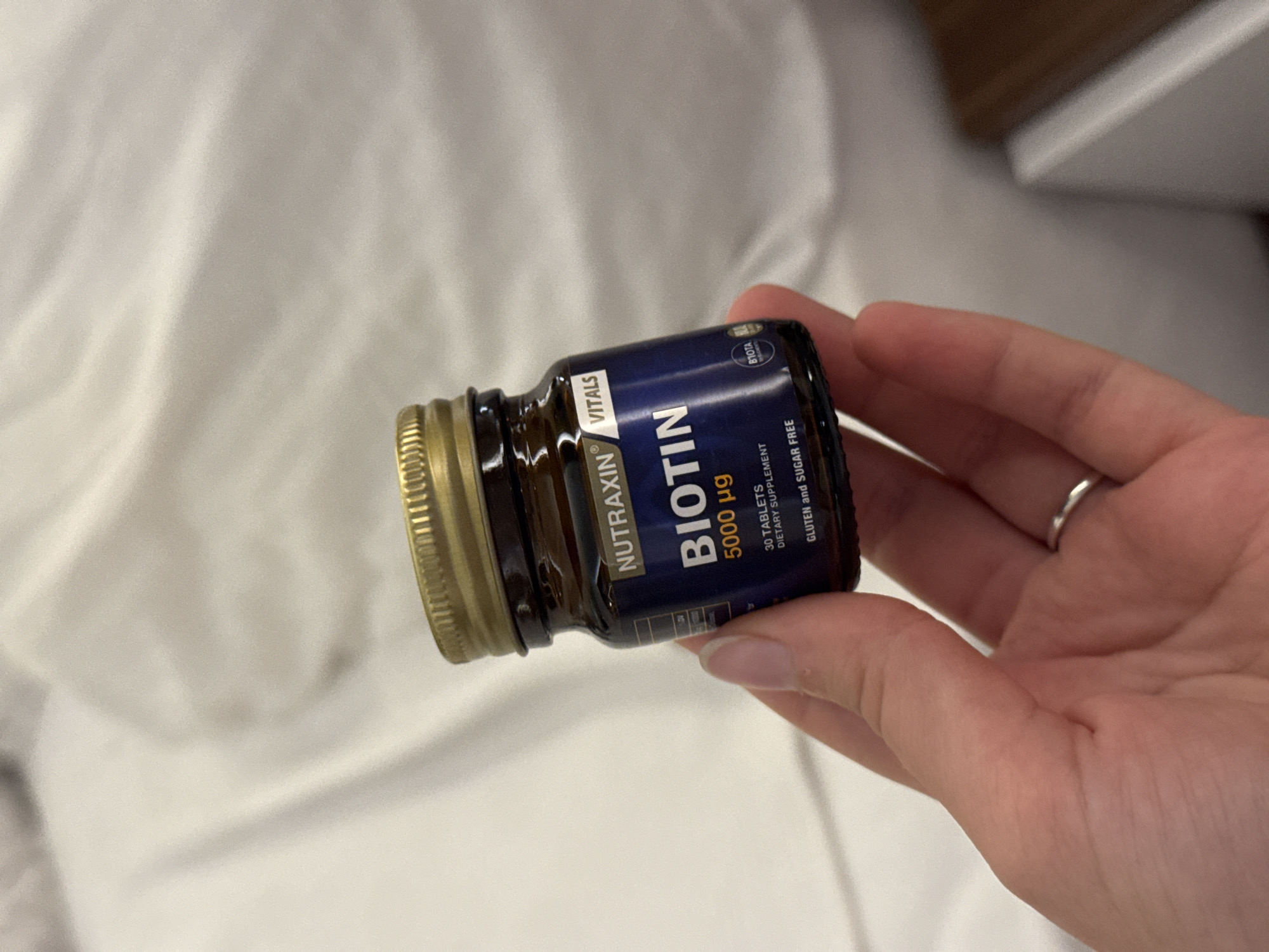 A hand is holding a small, dark brown glass bottle with a gold-colored cap. The bottle has a dark blue label with white text. The text on the label reads "NUTRAXIN VITALS BIOTIN 5000 µg 30 TABLETS DIETARY SUPPLEMENT GLUTEN AND SUGAR FREE". A small circular logo with "BIOTA" is visible on the right side of the label. The background is a blurred white surface. The image does not provide any information about the location in the city of Aksu, Turkey.
