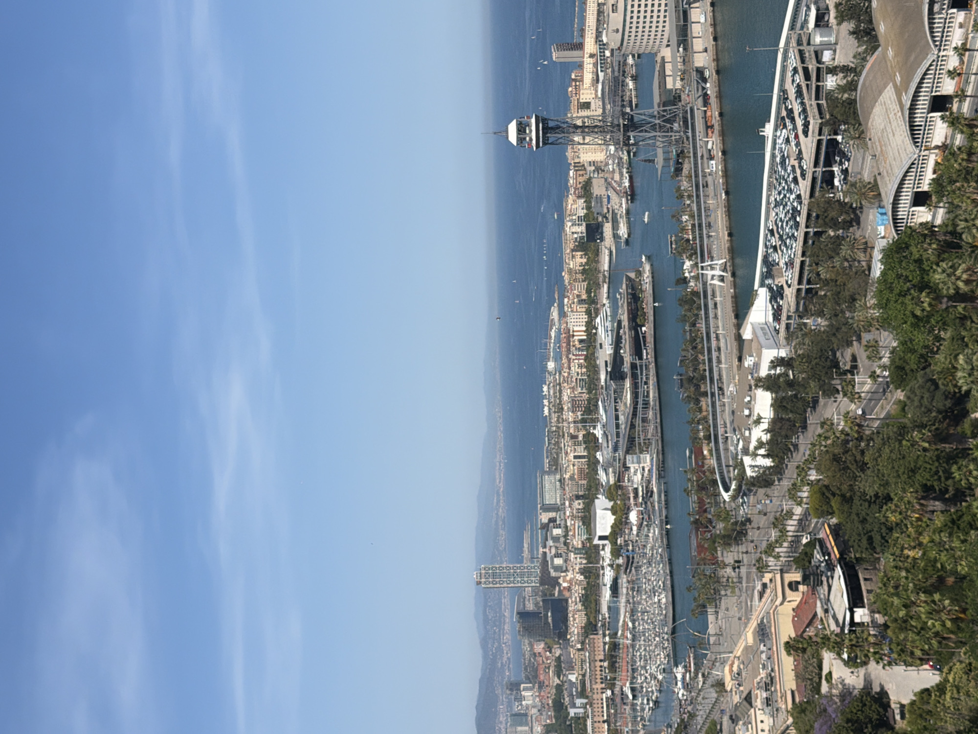 The image depicts a panoramic view of the Port of Barcelona, Spain, on a clear, sunny day. In the foreground, lush green trees and vegetation cover the lower portion of the frame, with some buildings nestled amongst them. A multi-story parking garage filled with numerous cars is prominently visible in the mid-ground, adjacent to the port's waterways. The harbor itself is teeming with a vast array of docked sailboats and yachts.
Further into the scene, several buildings of varying architectural styles line the waterfront, including modern high-rises and older structures. A distinctive telecommunications tower, characterized by its open lattice structure and a spherical observation deck at the top, stands tall overlooking the port. In the distance, the Mediterranean Sea stretches towards the horizon, dotted with numerous small sailboats. The city's skyline is visible in the background, with hills and mountains forming a backdrop to the urban landscape. The sky is a clear, vibrant blue with a few wispy clouds. No people are clearly discernible in the image.