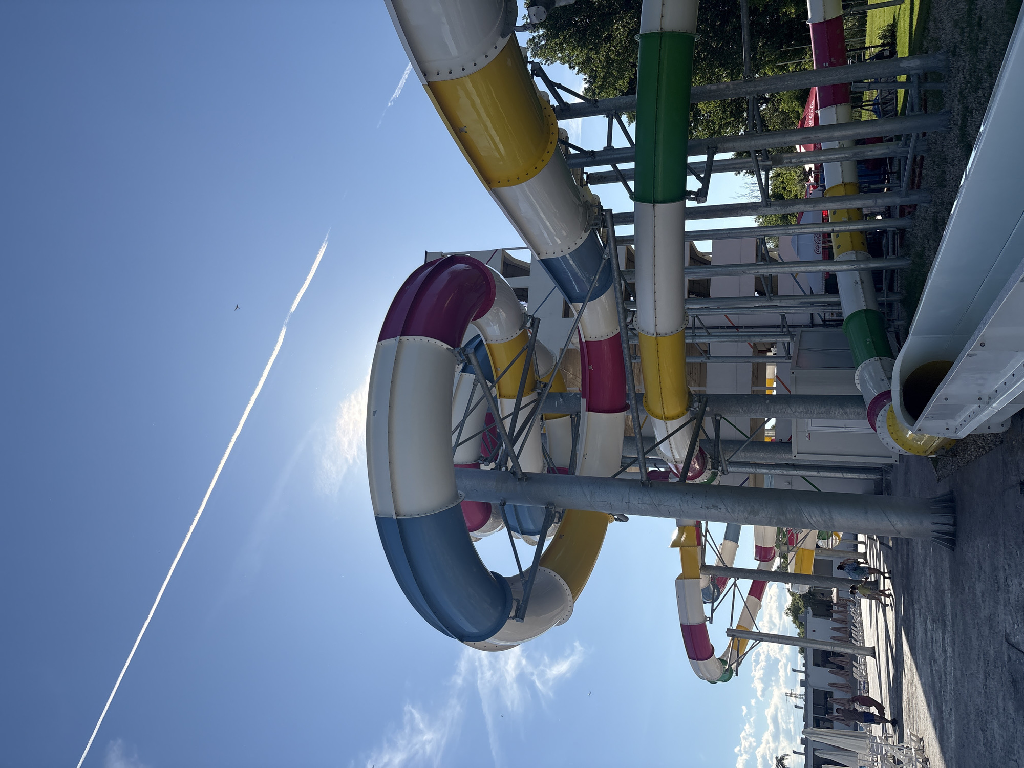 An eye-level, wide shot captures a water park with a bright blue sky overhead, featuring a prominent contrail left by an airplane and a small plane visible in the distance. The water slides are the main focus, constructed from cylindrical sections of various colors including white, blue, yellow, pink, and green, supported by a metal framework. The slides spiral and twist, creating a visually complex structure. Sunlight beams through gaps in the slides and between clouds, casting shadows on the paved ground below. In the foreground, a portion of a white slide is visible, leading away from the viewer. To the left, a walkway is lined with lounge chairs and umbrellas, with a few people walking in the middle ground. To the right, lush green trees and grass border the water park. The scene appears to be set in a sunny, recreational outdoor environment.