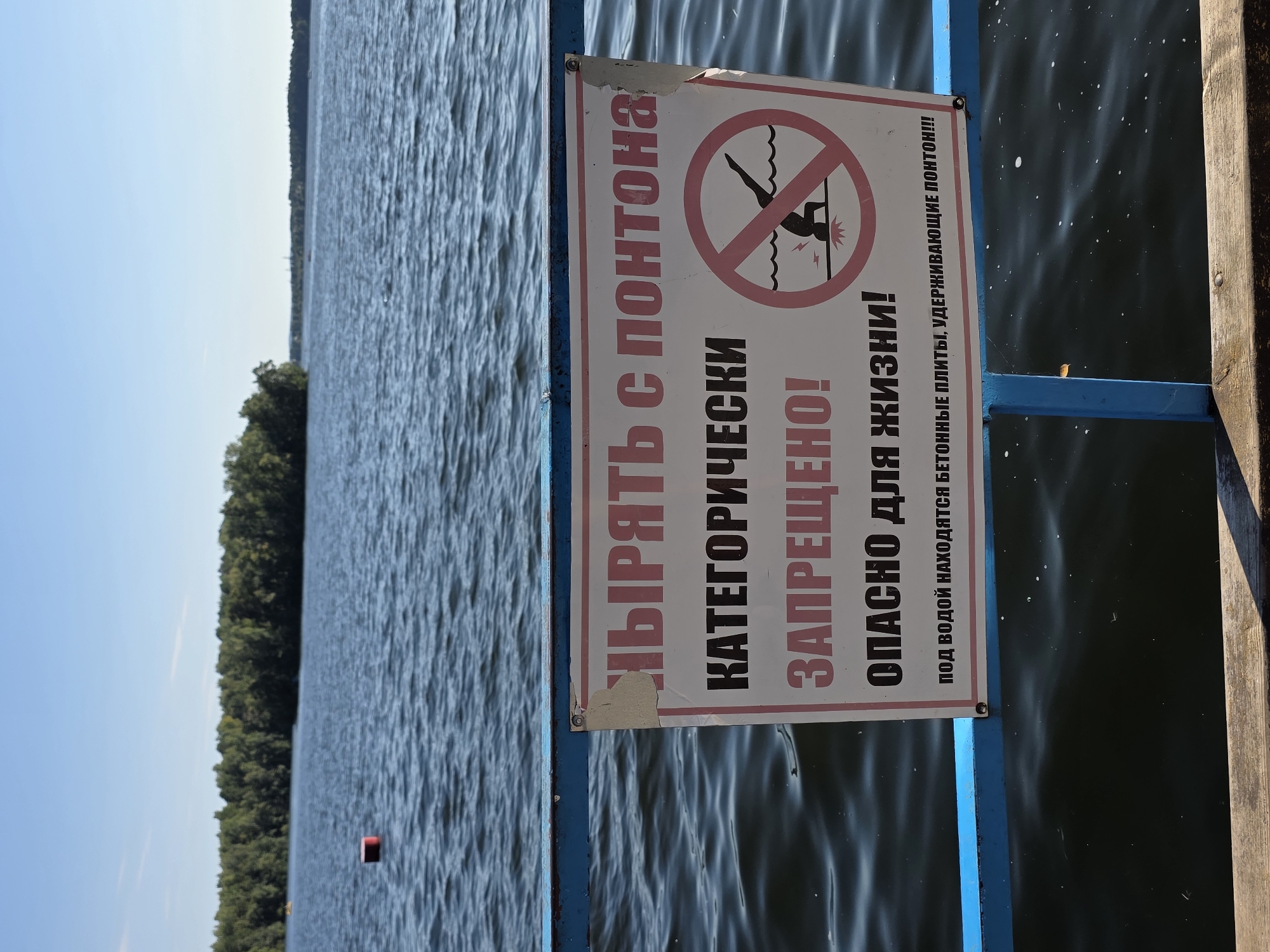 The image shows a sign on a blue railing overlooking a body of water. The sign is white with pink and black text and a prohibition symbol of a person diving. The text on the sign reads "JUMPING FROM THE PONTΟΟΝ IS STRICTLY PROHIBITED! IT IS DANGEROUS TO LIFE! UNDERWATER ARE CONCRETE SLABS HOLDING THE PONTOON!!!" The water is blue with ripples, and a distant treeline is visible on the horizon under a clear blue sky. A red buoy floats in the water. The railing is weathered and shows signs of rust. The scene appears to be an outdoor recreational area by a lake, possibly in Chebarkul, Russia, given the text on the sign is in Russian.