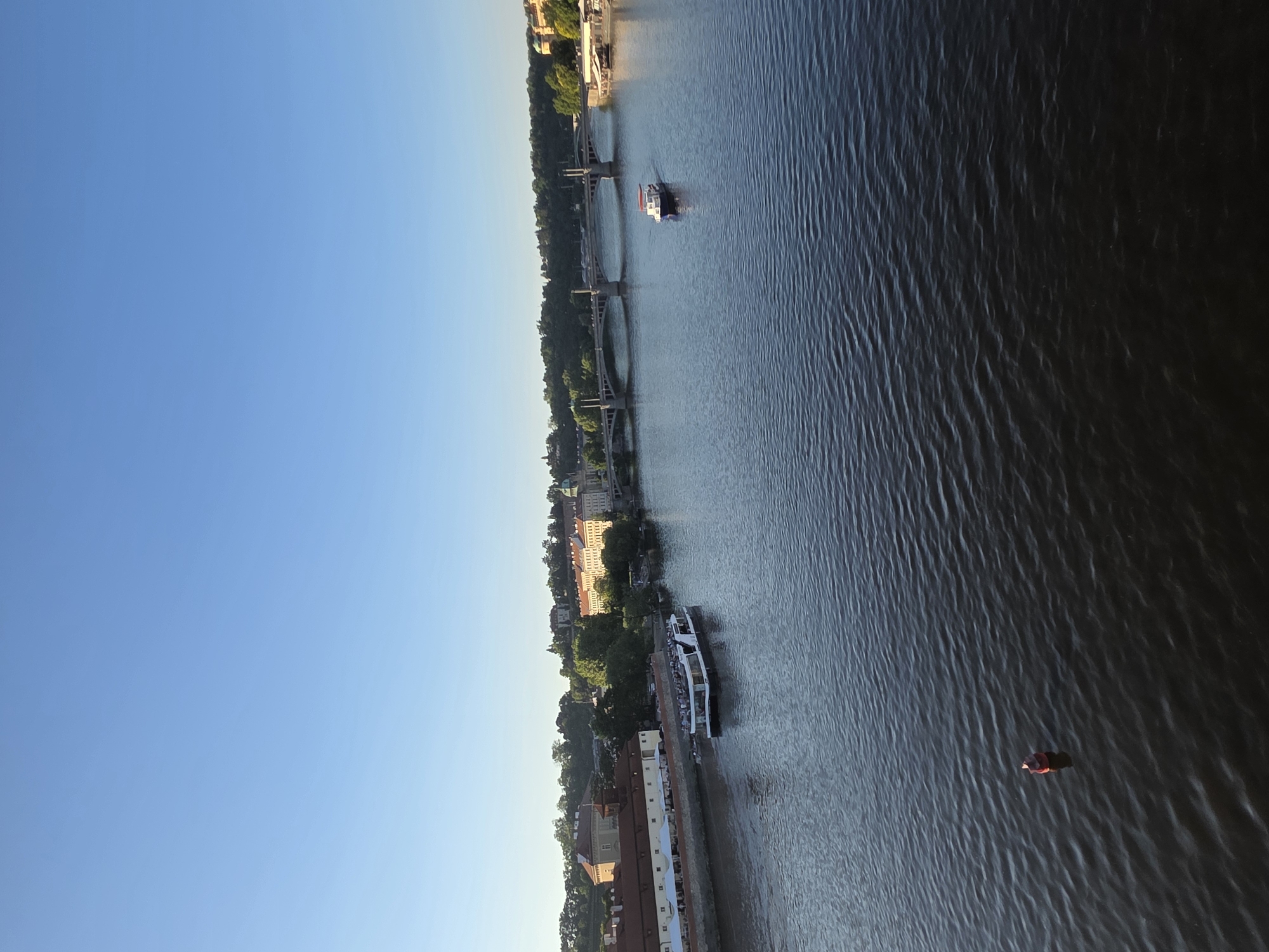 The image shows an aerial view of a river running through a city. The sky is clear and blue. Along the riverbank, there are buildings, trees, and infrastructure like bridges. Several boats are visible on the river. One boat is docked near a building. Another boat is mid-river, heading towards a bridge. A small, red-colored object is visible in the water. The location is in the city of Unknown, Unknown.