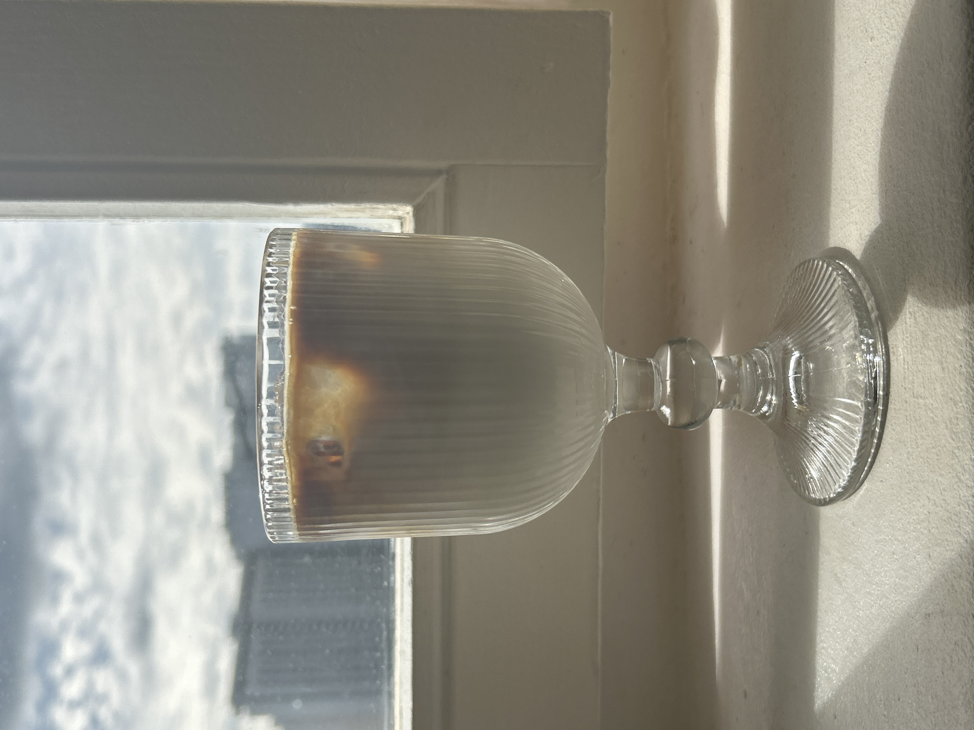 A glass of iced coffee sits on a windowsill. The glass is clear and ribbed, with a stem and a flared base. The coffee is layered, with a dark espresso on top and a lighter liquid, likely milk or cream, below. Ice cubes are visible in the coffee. Outside the window, a cloudy sky can be seen, with the blurry shapes of buildings in the distance. The scene is lit by natural light, casting shadows from the glass onto the windowsill. The location is not specified as Moscow, Russia, or any other specific city.