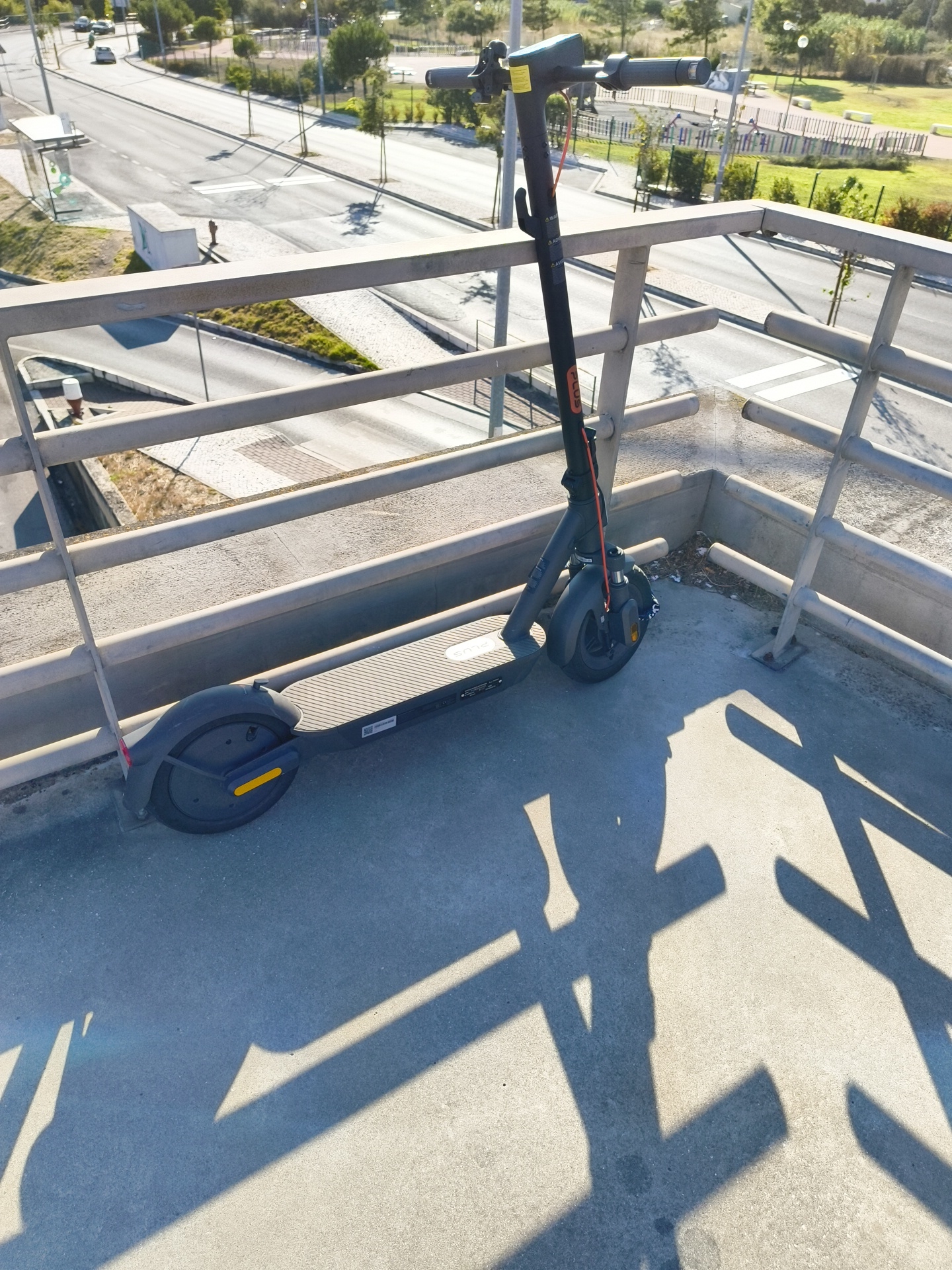 An electric scooter is parked on a concrete surface next to a metal railing. The scooter is dark gray with an orange charging cable attached and a yellow reflector on the rear wheel. The scooter's deck has a "plus" logo and some visible text and a QR code. The railing is light gray and runs along the edge of the concrete platform. Beyond the railing, a road is visible with cars driving on it, sidewalks, streetlights, and some green spaces with trees and a playground. The sun casts long shadows of the railing and the scooter onto the concrete surface. The scene appears to be in an urban or suburban environment. The location is identified as Cascais, Portugal.