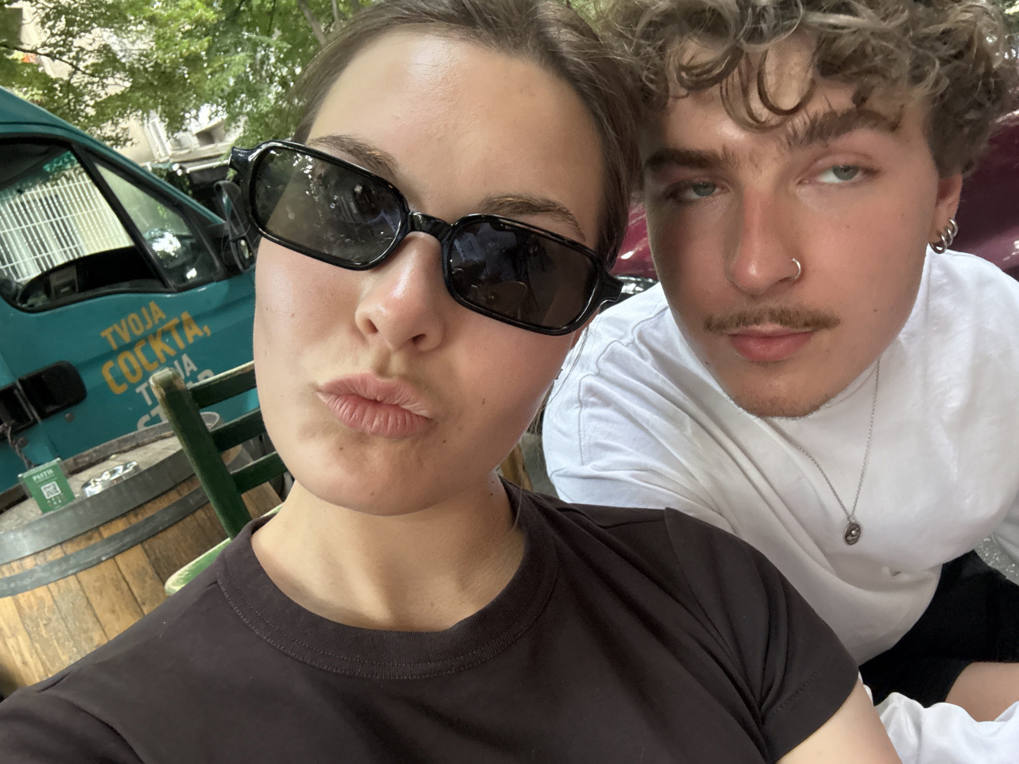 A close-up selfie captures two individuals, a woman and a man, in an outdoor setting in Belgrade, Serbia. The woman, positioned closer to the camera, wears black rectangular sunglasses and a dark brown t-shirt. She has a pursed-lip expression. Behind her, the man wears a white long-sleeved shirt, a silver necklace, and has a nose ring. The background reveals a teal van with the text "TVOJA COCKTA". They are seated at an outdoor café, as evidenced by the wooden barrel table and green metal chair. A small rectangular object, possibly a menu or advertisement, sits on the table. Green foliage is visible overhead.