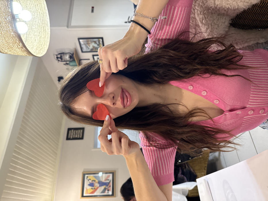A woman with long brown hair wears a pink and white striped sweater. She playfully holds red heart shapes over her eyes, sticking out her tongue. A gold ring adorns her finger. She wears a black bracelet, a silver bracelet and a silver necklace. Framed pictures hang on the wall in the background of a white room with a wicker lamp shade. The image is rotated.
