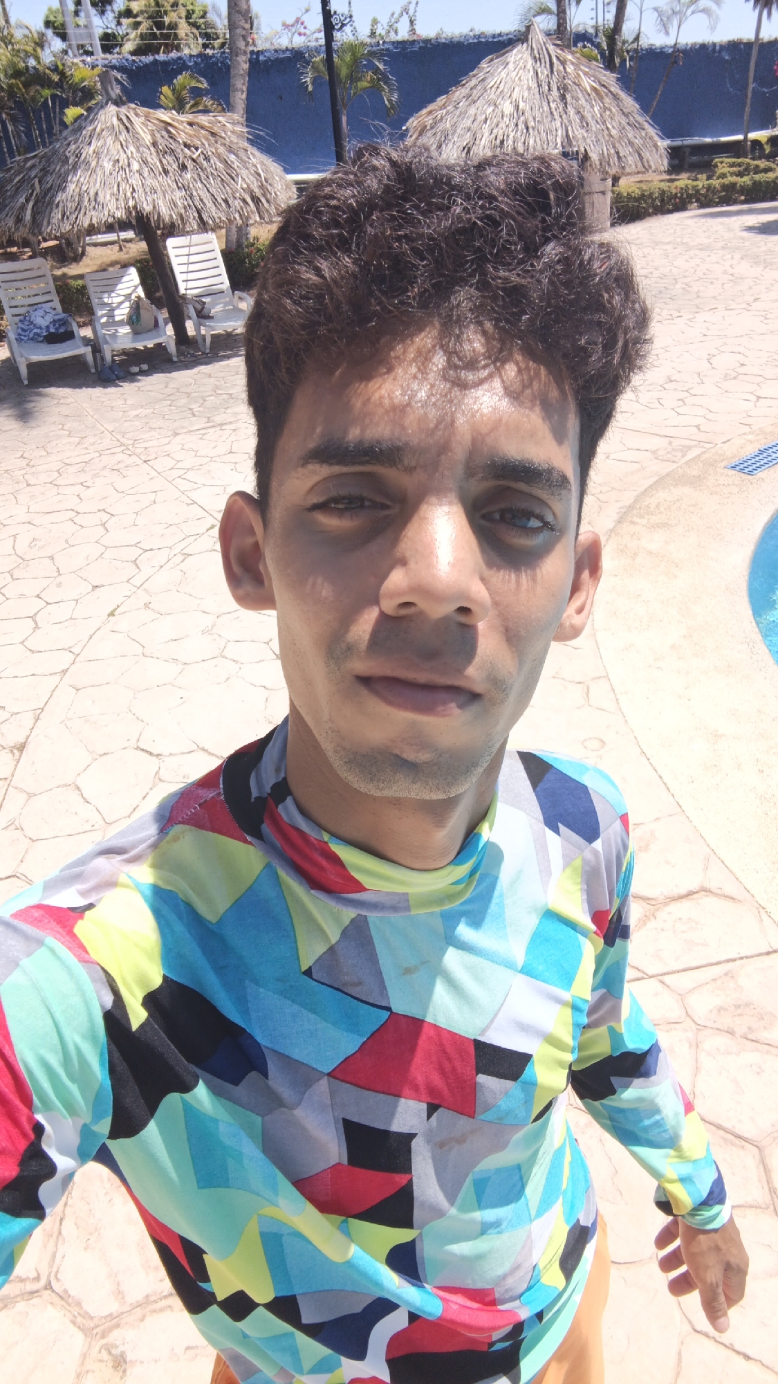 A young man takes a selfie outdoors on a bright, sunny day, likely at a resort or pool area. He has short, dark, curly hair and a clean-shaven or lightly stubbled face, looking directly into the camera with a neutral expression. He is wearing a long-sleeved shirt featuring a bold, multi-colored geometric pattern with shades of blue, green, yellow, red, black, and grey. His lower attire appears to be light orange or tan shorts.
The background shows a paved area with a distinctive cracked or tessellated pattern, leading to a bright blue swimming pool visible on the right side of the frame. In the distance, several thatched-roof umbrellas (palapas) shade white lounge chairs, suggesting a leisurely environment. Lush green vegetation, including palm trees, and a blue wall or fence are also visible further back, indicating a tropical setting. The strong, direct sunlight casts clear shadows, suggesting it is midday or early afternoon. The overall scene, with the resort-style amenities and tropical flora, is consistent with a location such as Playa El Agua, Venezuela. No visible text is present in the image.
