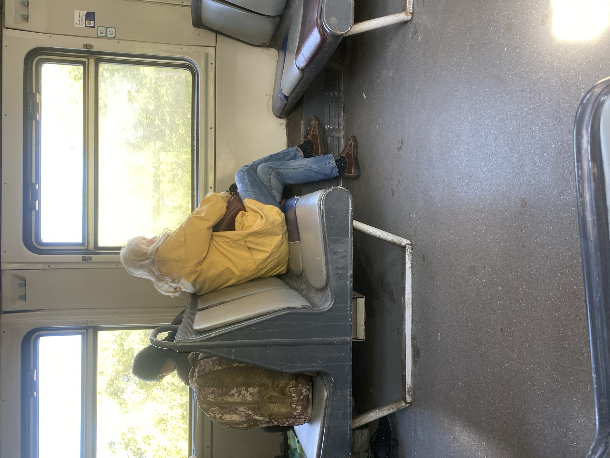 The image shows the interior of a train car. A person with long, white hair is lying across two seats, wearing a yellow jacket, blue jeans, brown shoes, and black socks with white dots. A person is sitting facing them on the lower seats, wearing a dark-colored shirt or jacket and with a camouflage-patterned bag. There are windows to the outside that reveal trees. The location is not able to be verified as Matiashivka, Ukraine.