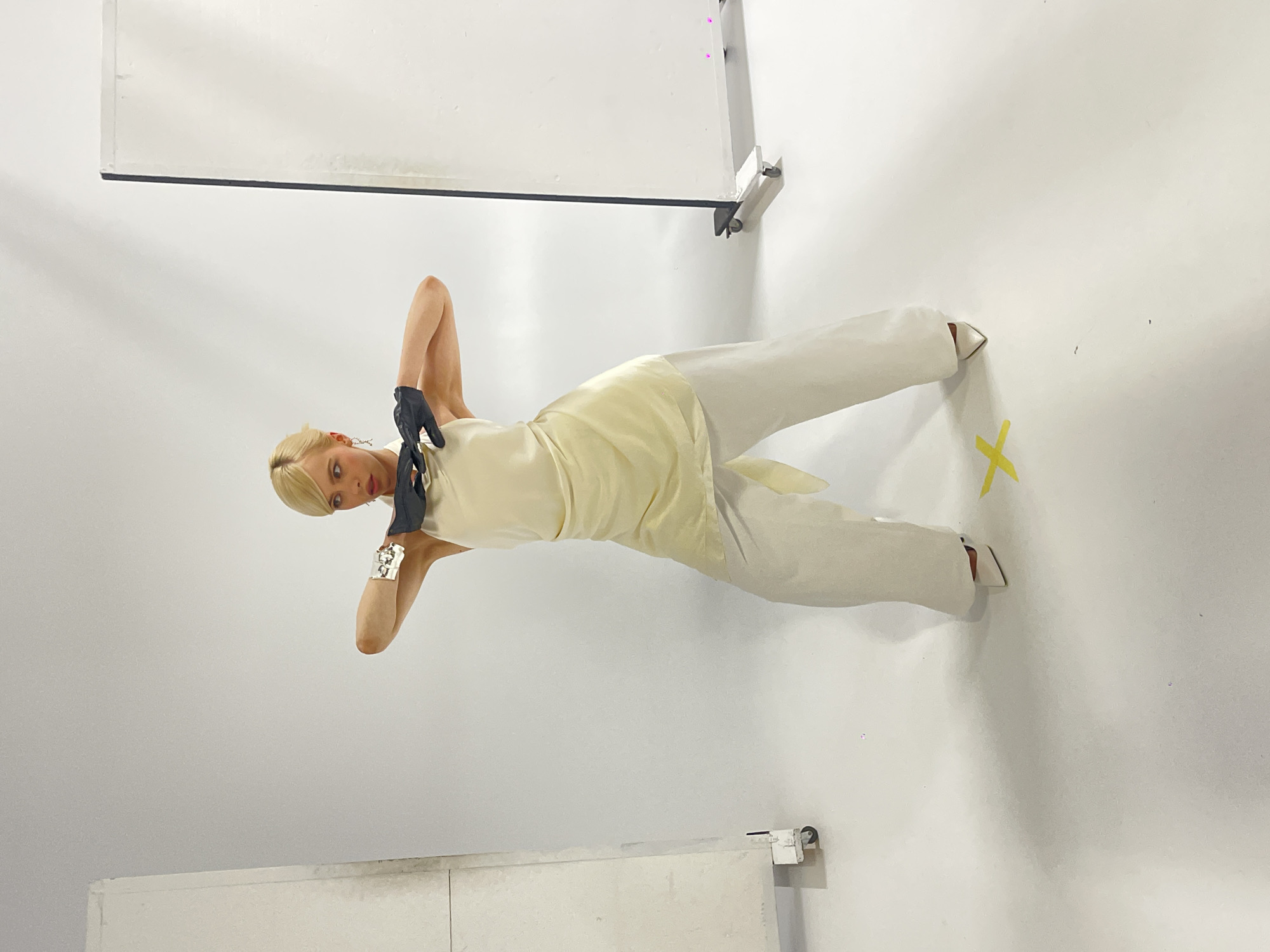A person with blonde hair is posed in a studio with white walls. They are wearing a pale yellow, sleeveless top with a sash tied at the waist and matching wide-leg pants. Black gloves are worn on both hands. A large, silver, cuff-like bracelet is visible on the left wrist. The person's head is tilted, and their eyes are looking towards the viewer with a neutral expression. They are positioned with their body angled, and one arm is raised, bent at the elbow, with the hand near their head. The other arm is bent and extended downwards, with the hand resting on their leg. A yellow "X" shaped sticker is visible on the wall behind them, along with a white horizontal structure at the bottom of the frame. The lighting suggests an indoor studio environment.