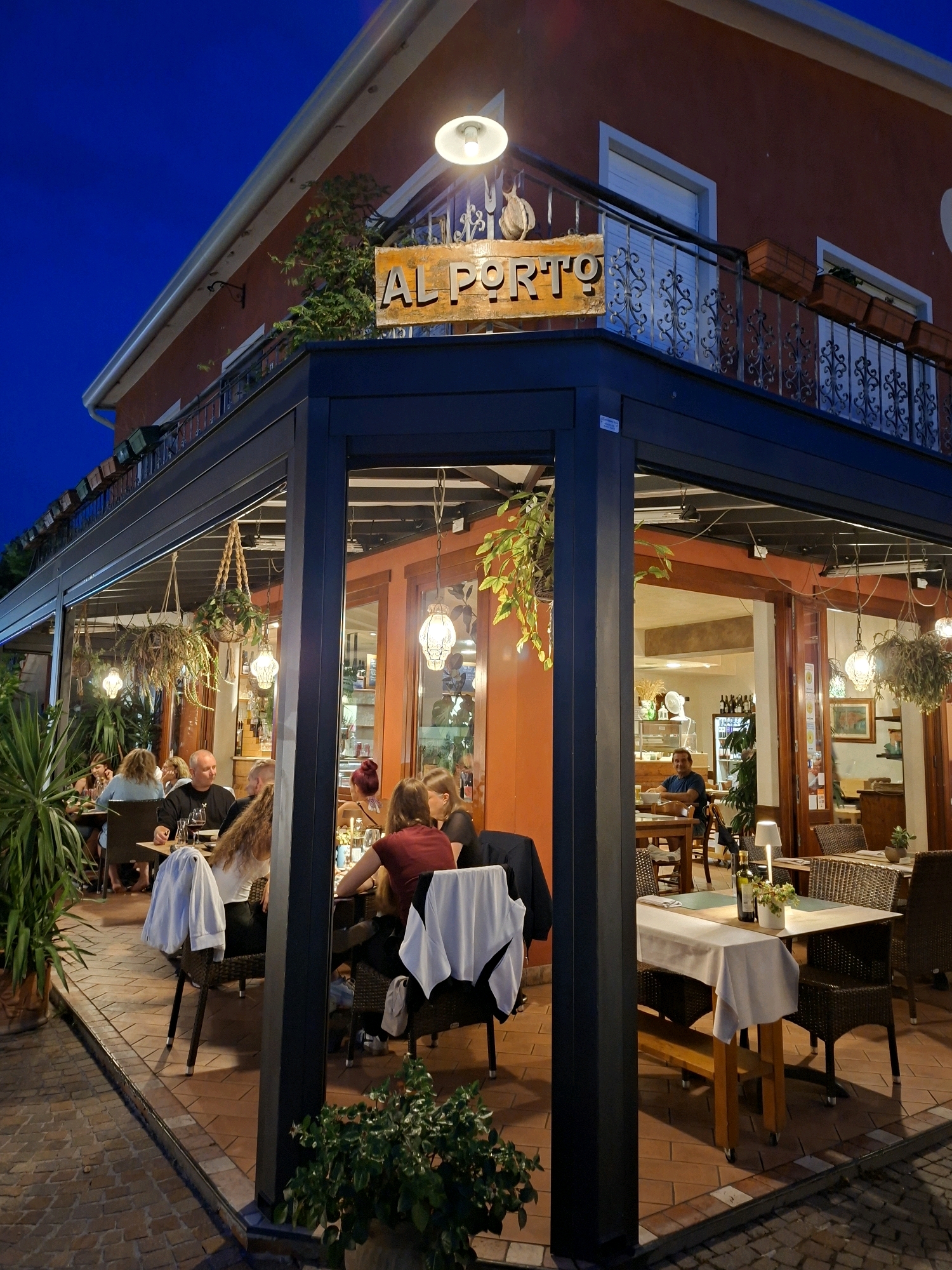 The image captures an outdoor seating area of a restaurant named "AL PORTO" at dusk in Caorle, Italy. The restaurant has a prominent red facade and a covered seating area with a glass enclosure. Inside the covered area, several tables are occupied by diners. To the left, a group of people are seated at a table, some facing away from the camera. To the right, another table is set with white tablecloths and place settings, with a few individuals seated, one looking towards the camera. Hanging plants and decorative lights add to the ambiance. Beyond the seating area, the interior of the restaurant is visible, with shelves displaying bottles and a man sitting at a table. The ground is paved with bricks, and various potted plants surround the seating area. The sky above is a deep blue, indicating the late hour.