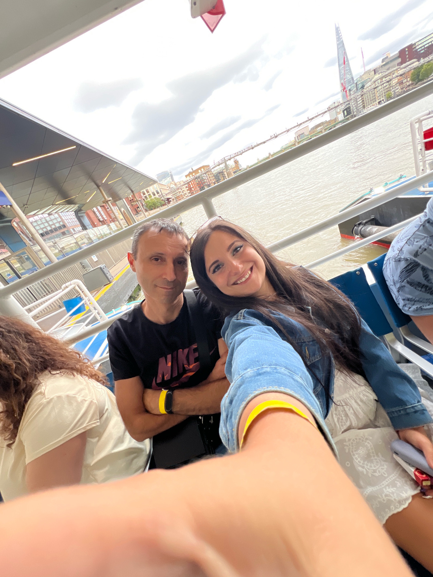 A selfie is taken from the perspective of a person's arm, showing a man and a woman sitting on a boat in London. The man, wearing a black t-shirt with a "Nike" logo, has his arms crossed. The woman, wearing a denim jacket and a white lace dress, is smiling at the camera. In the background, the River Thames is visible, along with buildings and the Shard skyscraper. The boat appears to be at a dock or pier, with railing and seating for passengers. The sky is overcast.