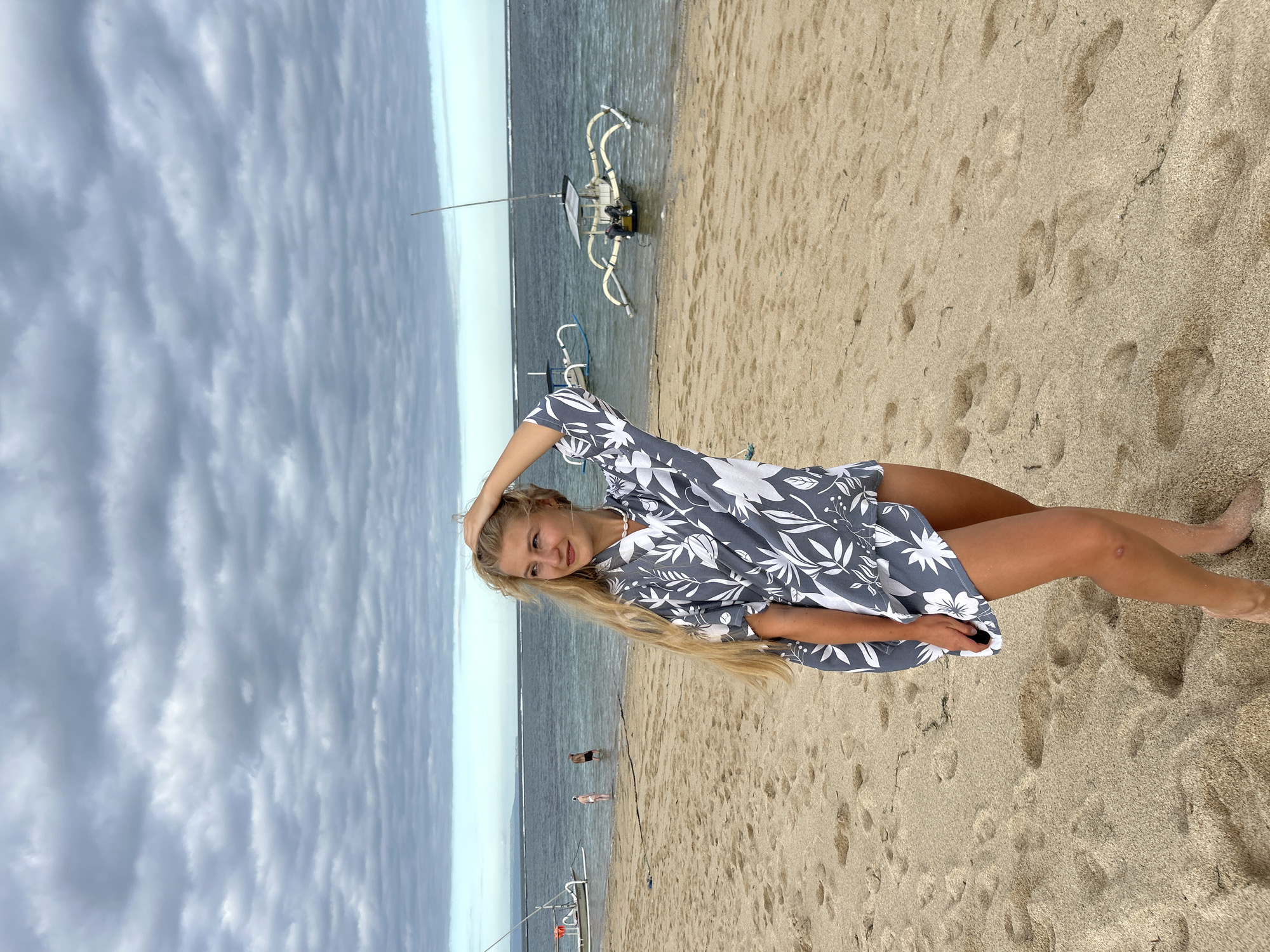 The image depicts a young woman lying on a sandy beach. She has long blonde hair and is wearing a grey and white floral-patterned dress. Her right hand is positioned behind her head, and she is looking towards the camera.
The beach is characterized by tan-colored sand with numerous footprints. The ocean is visible in the background, with some small waves. There are two people standing on the shoreline. A traditional outrigger boat is docked in the water. The sky is overcast with grey clouds.
The location is Kuta Selatan, Indonesia.