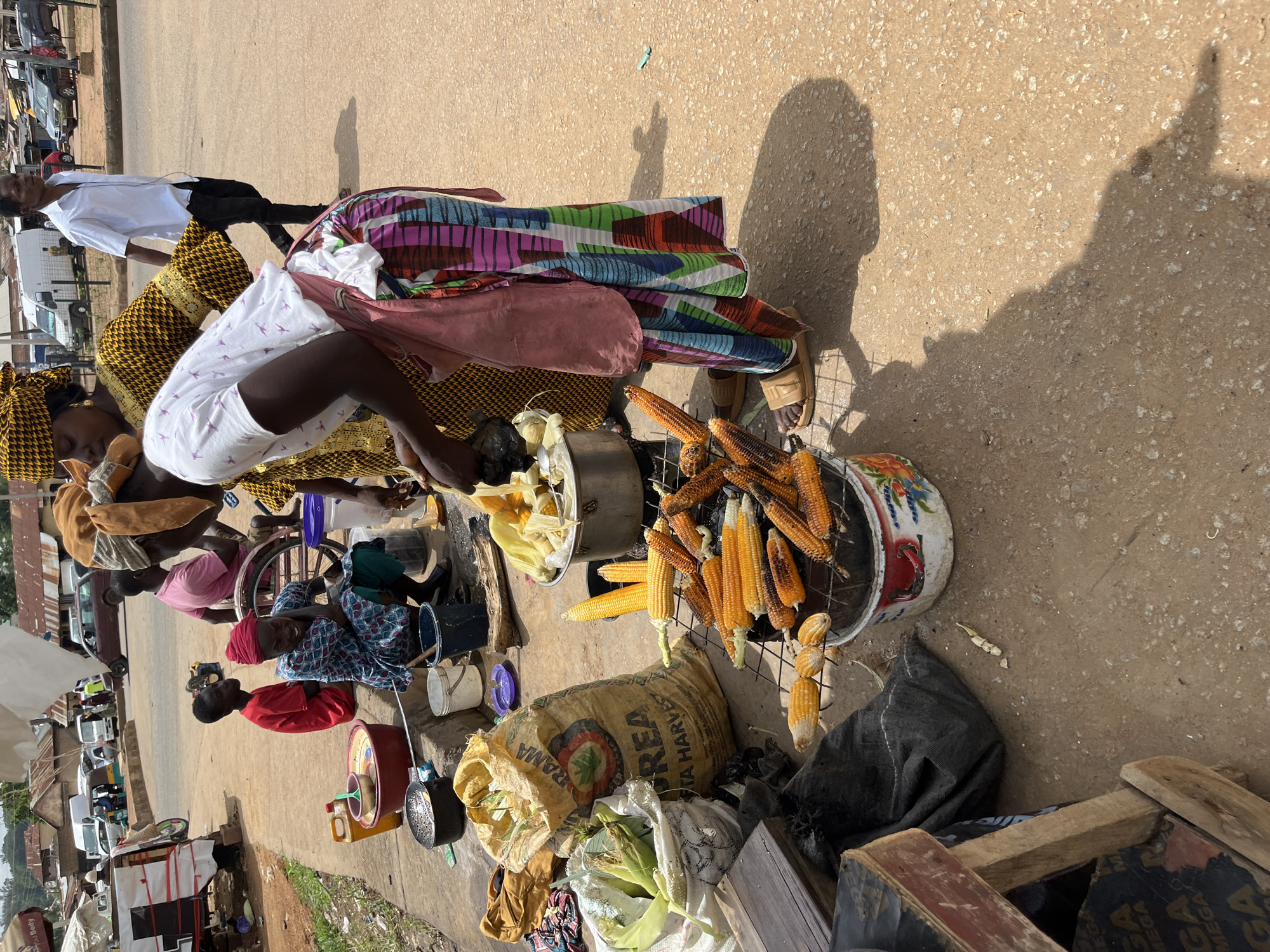 In Bukuru, Nigeria, a street vendor is roasting corn on a grill. The vendor, a woman wearing a colorful patterned skirt and a pink apron, is standing behind the grill, attending to the corn. Several cobs of corn are already cooked and arranged on the grill, some appearing slightly charred. In front of the grill, there is a large sack of corn and another bag containing husked corn. A large pot, possibly used for boiling corn, sits nearby. Other individuals are visible in the background, including a man in a white shirt walking past and a woman in a yellow patterned dress. The scene takes place on a dusty street with a few vehicles and buildings visible in the distance, suggesting a market or busy roadside area. The overall impression is of a vibrant street-side food vending operation.