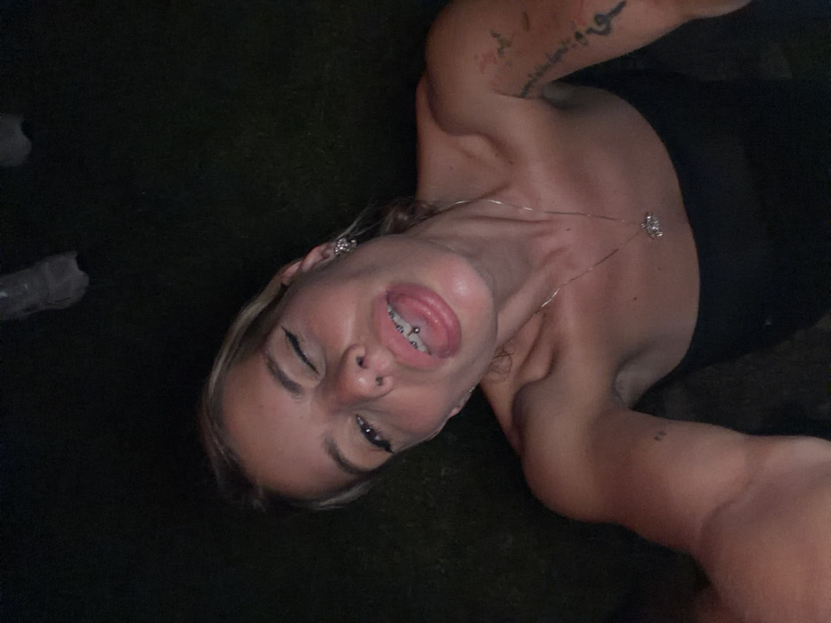 The image depicts a fair-skinned individual with blonde hair, captured in an upward-angled selfie at night or in a dimly lit environment in the city of Unknown, Indonesia. The subject's left eye is closed in a wink, while the right eye remains open; both eyes feature prominent black eyeliner. A stud piercing is visible on the right nostril, and a barbell piercing is present on the extended tongue. Dental braces are visible on the upper teeth. The individual wears a strapless black top, silver-colored hoop earrings, and a silver-colored necklace with a small, light-colored pendant. Tattoos are present on both arms: script or an abstract design on the right arm and a small dark dot on the left bicep. The background is predominantly dark, showing blurred green textures consistent with grass or foliage and two indistinct, light-colored objects in the upper frame, possibly plastic bottles.