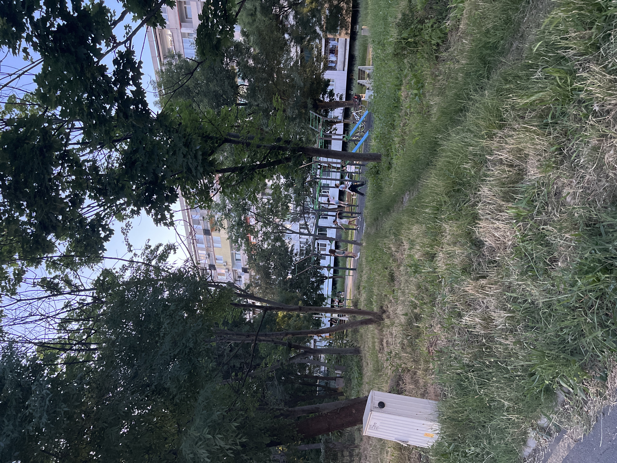 The image depicts a residential courtyard with a playground, located in Burgas, Bulgaria. In the foreground, tall, dry grass lines a dirt path that winds towards the center of the frame. To the left, a white electrical box stands by the roadside. Beyond the grass, a playground with green and blue structures, including a slide and monkey bars, is visible. A bicycle is parked near the playground. Several individuals, appearing to be children and adults, are present on and around the playground equipment. In the background, a multi-story residential building with light-colored facades and numerous windows dominates the skyline. Trees with dense green foliage are interspersed throughout the scene, creating a dappled light effect. The sky is clear and blue, suggesting it is daytime.