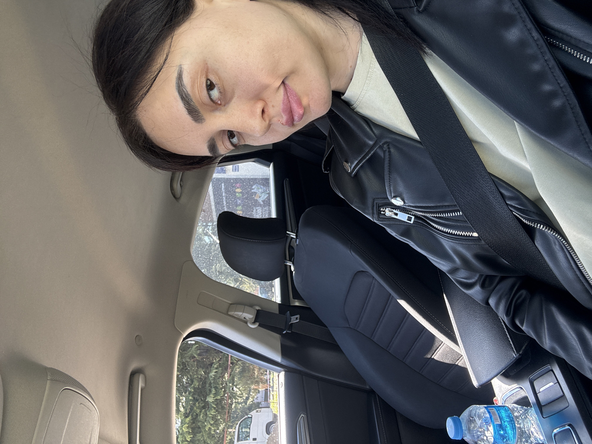 A woman is seated in the passenger seat of a car. She is wearing a black leather jacket over a light-colored shirt, and a seatbelt is fastened across her body. The interior of the car includes black leather seats, a water bottle in the console, and glimpses of trees and buildings visible through the car windows. The location is possibly Tbilisi, Georgia.