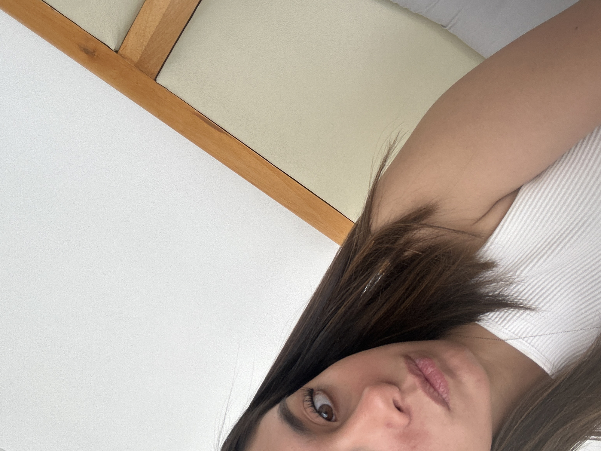 A close-up, tilted shot features a portion of a person's face (an eye, nose, and part of the mouth) in the lower-right quadrant. The person has brown hair and appears to be wearing a white, ribbed shirt or top. The background consists of white wall and a wooden frame or furniture element with a beige or light-colored cushion/surface attached. There is no information about the location of the city of Unknown, Unknown.
