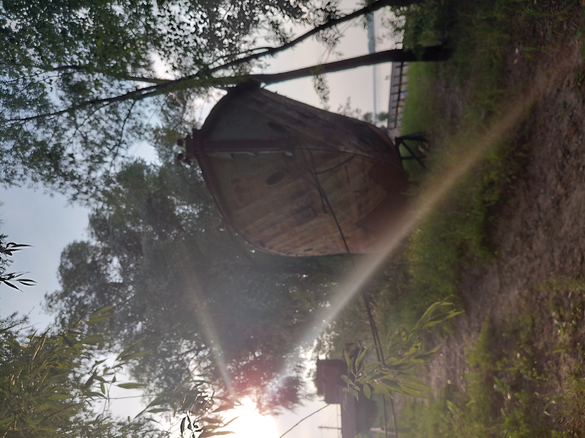 The image shows a rusted, disused boat on land, situated amidst trees and vegetation. The sun is visible and casting rays of light across the scene. In the background, a body of water and a distant shoreline can be seen. There are no people or animals present. The boat appears to be an old, possibly abandoned, vessel. The overall impression is one of decay and neglect in a natural setting.
This image was taken on the bank of the Ob River in the Sovetsky District of Novosibirsk, Russia. The area is known for its natural beauty and recreational opportunities, despite being part of a large urban area. The presence of the abandoned boat suggests a history of maritime activity or perhaps a discarded piece of industrial equipment. The overgrown vegetation and sun glare contribute to a somewhat melancholic atmosphere.