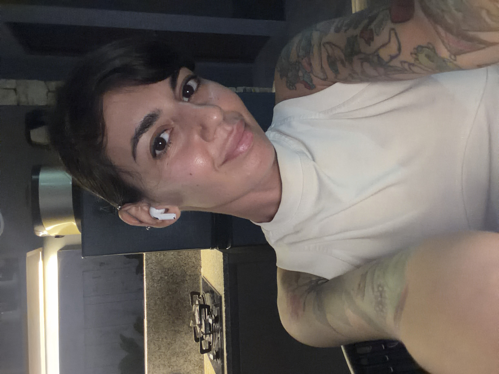 An adult female takes a close-up, chest-up selfie indoors, likely in a modern kitchen or open-plan living area, in Kuta Selatan, Indonesia. She has dark, short hair styled with bangs, dark brown eyes, and fair skin with a subtle sheen. Her expression is a slight, closed-mouth smile. She wears a light-colored (off-white or cream) high-necked sleeveless top. A white wireless earbud is placed in her right ear, which also features a silver-colored bar piercing on the upper cartilage. Both of her arms are adorned with colorful, full-sleeve tattoos featuring abstract and cartoon-like designs.
The background reveals a dark-themed kitchen area. On the left, a black gas stovetop with two burners and silver grates is visible on a light-colored, speckled countertop. Dark-colored base cabinets extend beneath the counter. A horizontal strip light mounted under an upper cabinet illuminates the countertop. To the right, behind the subject's head, dark wall units or cabinets are present, with a cylindrical appliance featuring a metallic gold-toned lid visible on top of one. Reflections on the dark surfaces indicate a glossy finish. The overall lighting is artificial, suggesting an evening or nighttime setting.