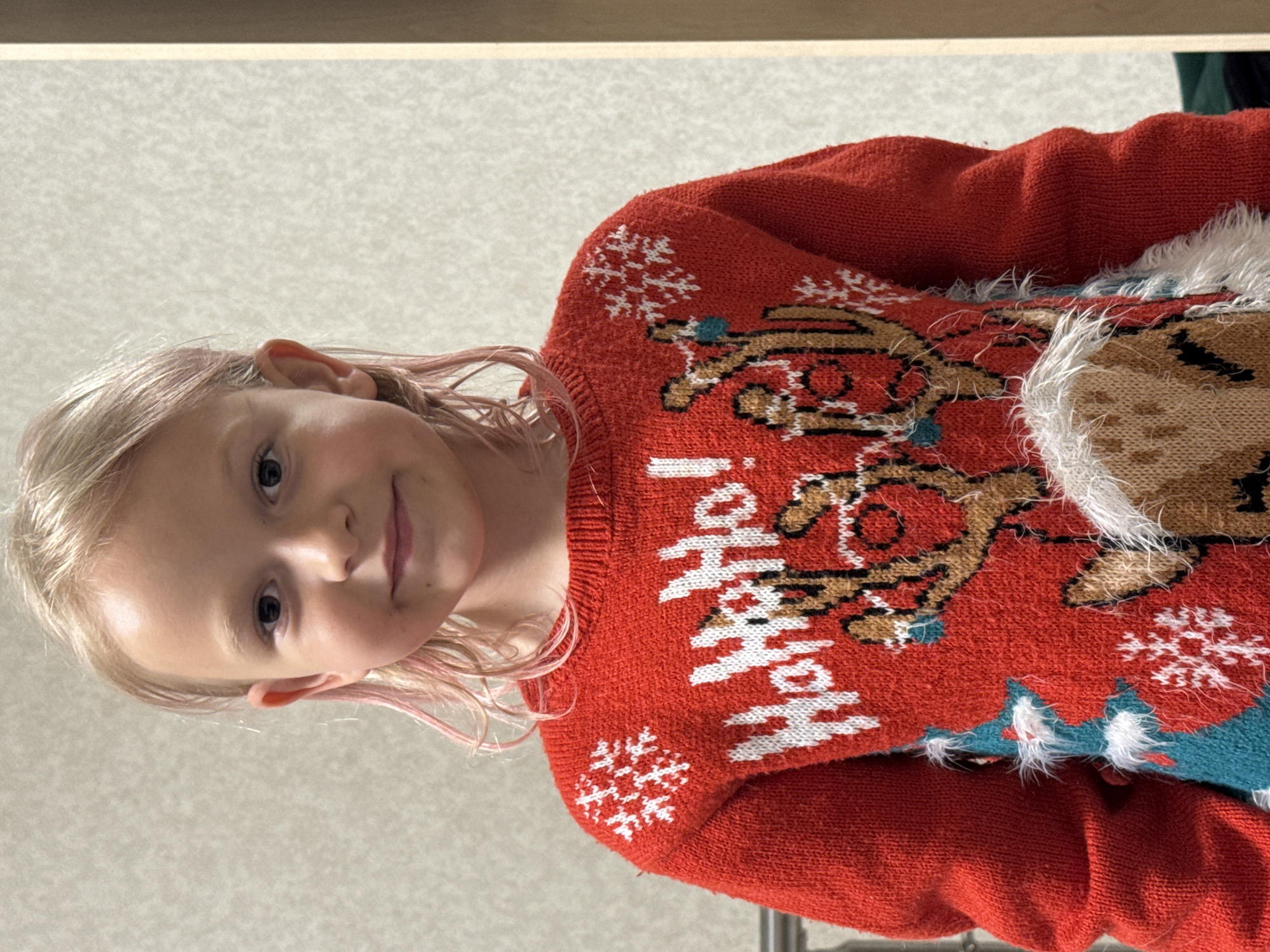 A young girl with blonde hair, tinged with pink, stands in front of a textured wall. She is wearing a red, long-sleeved sweater adorned with a Christmas theme, featuring white snowflakes, the phrase "HoHoHo!" in white lettering, and a reindeer design with fuzzy white accents. The girl's gaze is directed at the camera, and she has a subtle smile. The lighting suggests an indoor setting, possibly a room with a neutral wall and a wooden frame visible on the right side of the image.