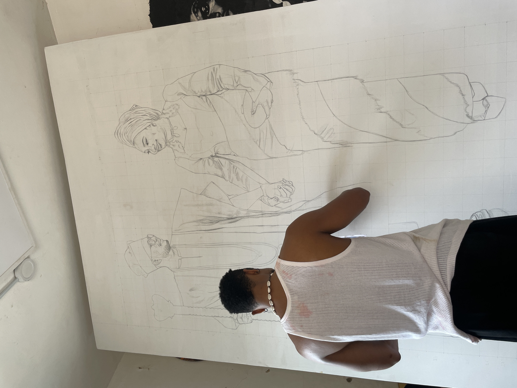 The image shows a person standing in front of a large canvas on a wall, drawing. The canvas is white and appears to have a grid drawn on it. There is a drawing on the canvas of two people. The person in the foreground is wearing a white tank top and black pants, with a necklace made of what appears to be seashells. The background is a white wall with a painting or poster visible at the top of the frame. The location is unknown.