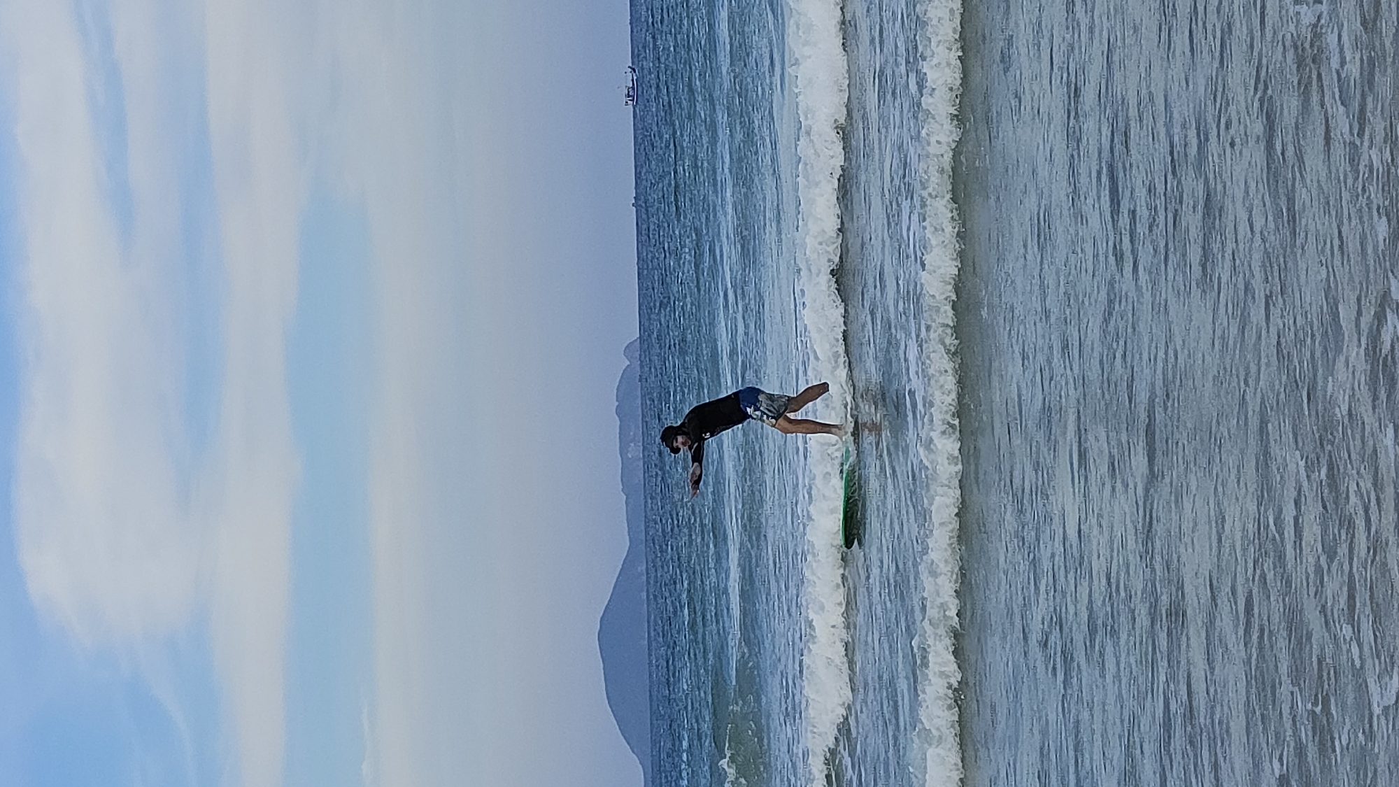 A man in dark attire and patterned shorts is surfing on a green board in the ocean at Bai Dai Beach, Cam Lâm, Vietnam. The sea has small white-capped waves and the shoreline extends towards the right edge of the image. In the background, a mountain silhouette can be observed along the coastline. The sky is partially covered by clouds on the left side of the image, transitioning into a clear sky above the ocean. A ship is visible in the distance at the top.
