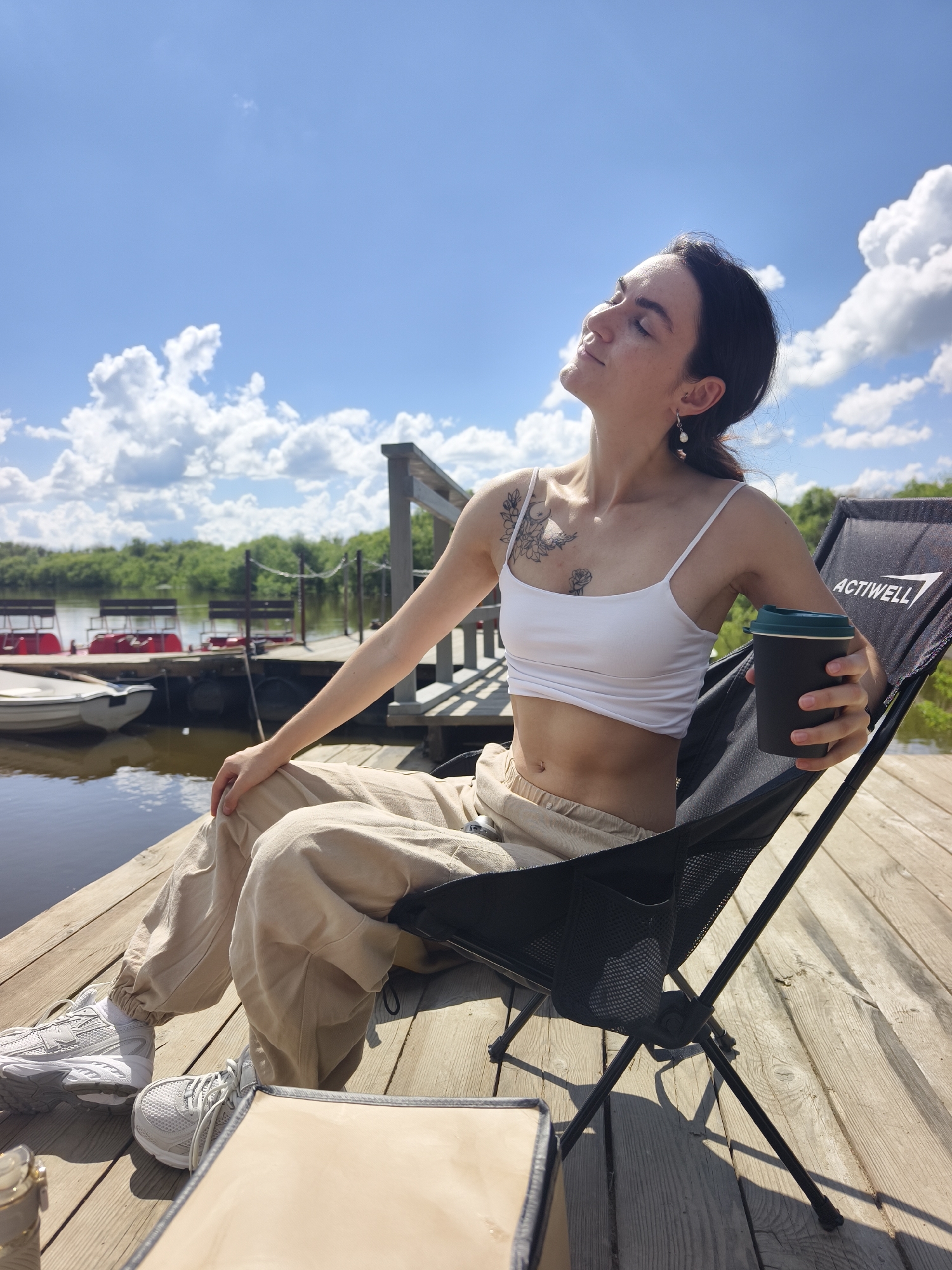 A young woman with dark hair, wearing a white crop top, light-colored sweatpants, and white sneakers, sits in a black folding chair on a wooden pier. She is holding a dark-colored cup with a green lid. Her eyes are closed, and her face is tilted towards the sun. She has a tattoo on her chest. Behind her, a body of water is visible, with several pedal boats docked and lush green trees in the background under a bright blue sky with scattered white clouds. The pier is made of light-colored wooden planks. The scene appears to be in the city of Onokhino, Russia, likely a recreational area by a river or lake.