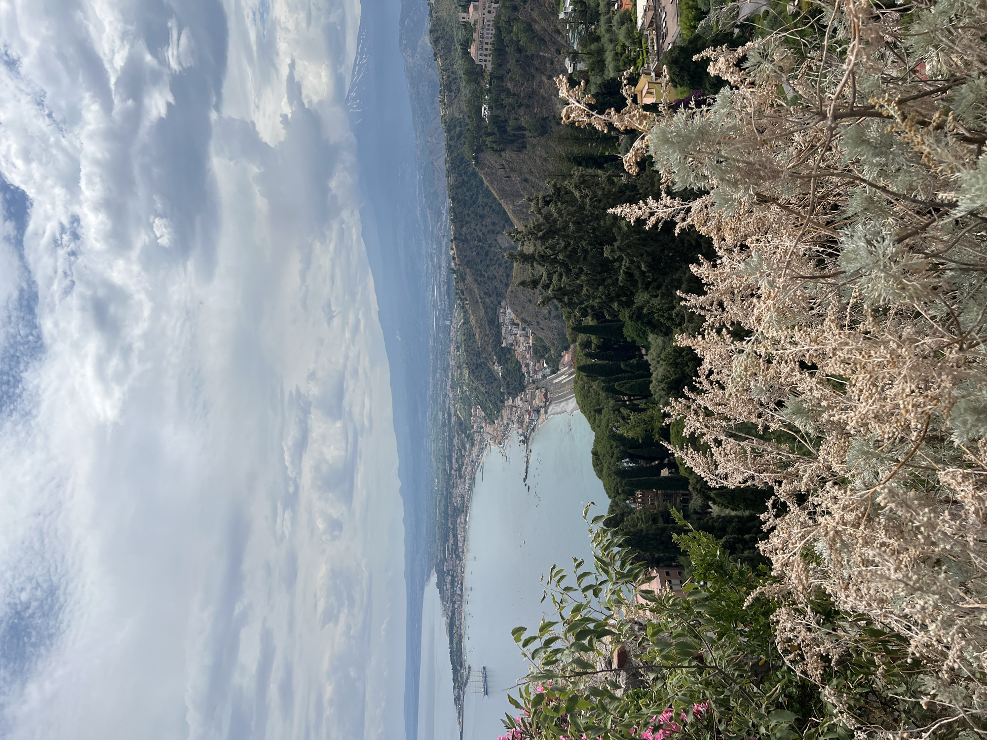 The image is a high-angle panoramic view of Taormina, Italy, taken on a cloudy day. In the foreground, there are dry, light-colored plants and some green foliage, with pink flowers visible on the left. The middle ground shows the coastal town of Taormina with its buildings stretching along the coastline and up the hills. A large, multi-masted sailing ship is anchored in the calm, blue sea, alongside numerous smaller boats. The background features a chain of mountains, with Mount Etna prominently visible, its peak capped with snow and a plume of smoke rising from it. The sky is filled with dramatic, heavy clouds. The scene captures a scenic vista of the town, sea, and volcano.