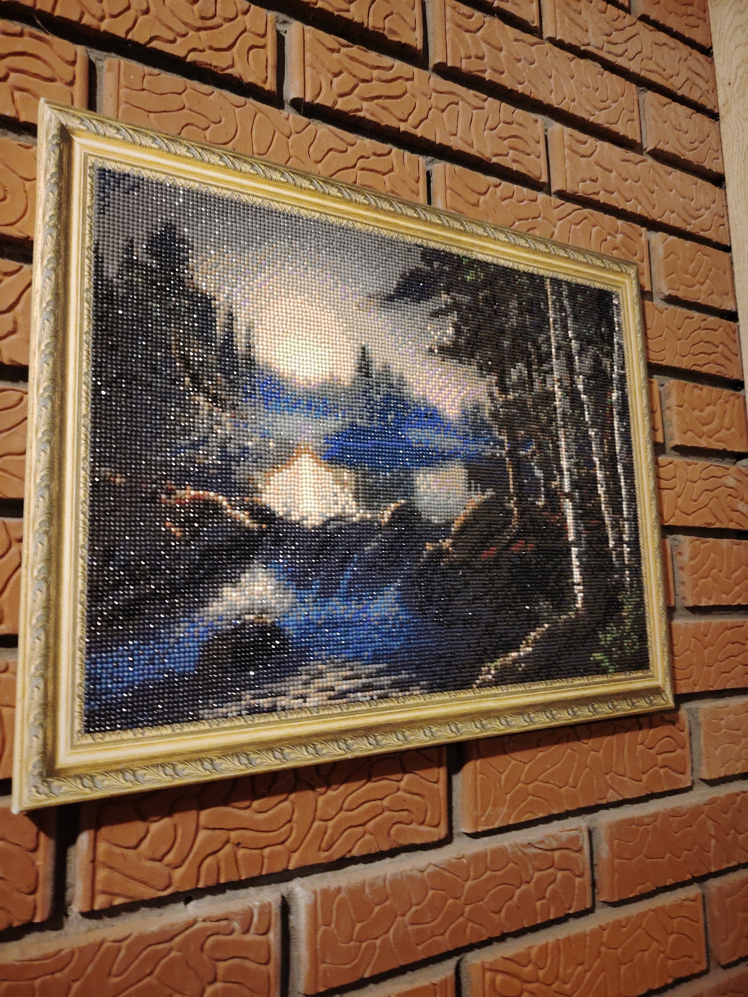 A framed, rectangular artwork hangs on a brick wall. The artwork depicts a landscape with trees, a body of water, and a light-filled sky. The artwork appears to be created using small, sparkling elements, possibly beads or crystals. The frame is gold-colored and ornate. The brick wall is textured and orange-brown.