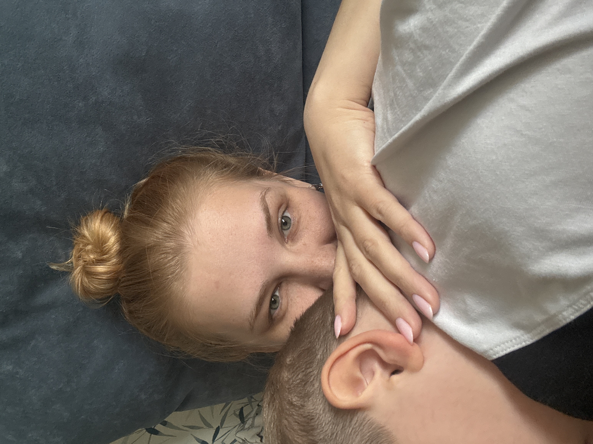 A woman with red hair in a bun is lying down. Her head is resting on a blue cushion. Her right hand is near a person’s face, their head partially visible, with their right ear clearly seen. The other person has short, light-brown hair. A gray garment is draped over part of the scene, possibly obscuring more of the second person. The background appears to be an interior space with a dark couch or bed. The location is not discernible from the image.