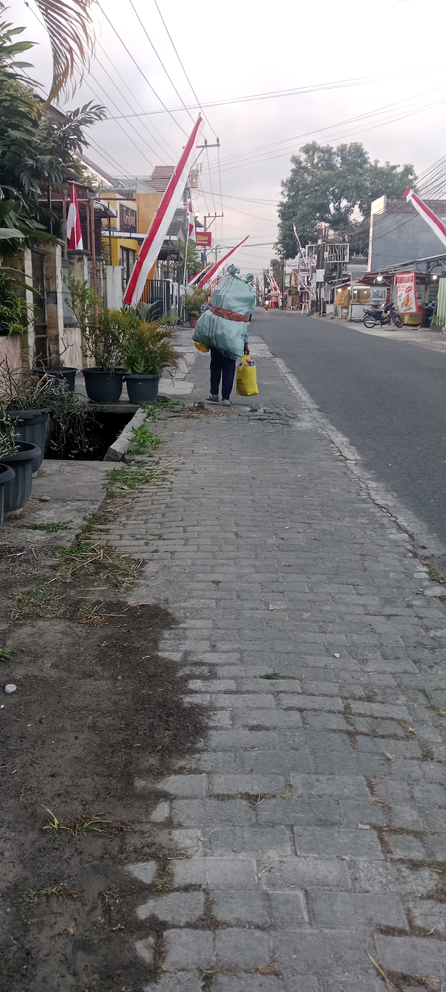 A person walks along a brick sidewalk beside a street in Maguwoharjo, Indonesia. The person is carrying a large, light blue sack over their left shoulder and a yellow bag in their right hand. They are wearing dark pants and light-colored shoes. The street is lined with buildings and small shops, some displaying red and white Indonesian flags. Trees and power lines are visible in the background. The sky is overcast. The sidewalk is bordered by potted plants on the left side and the street on the right.