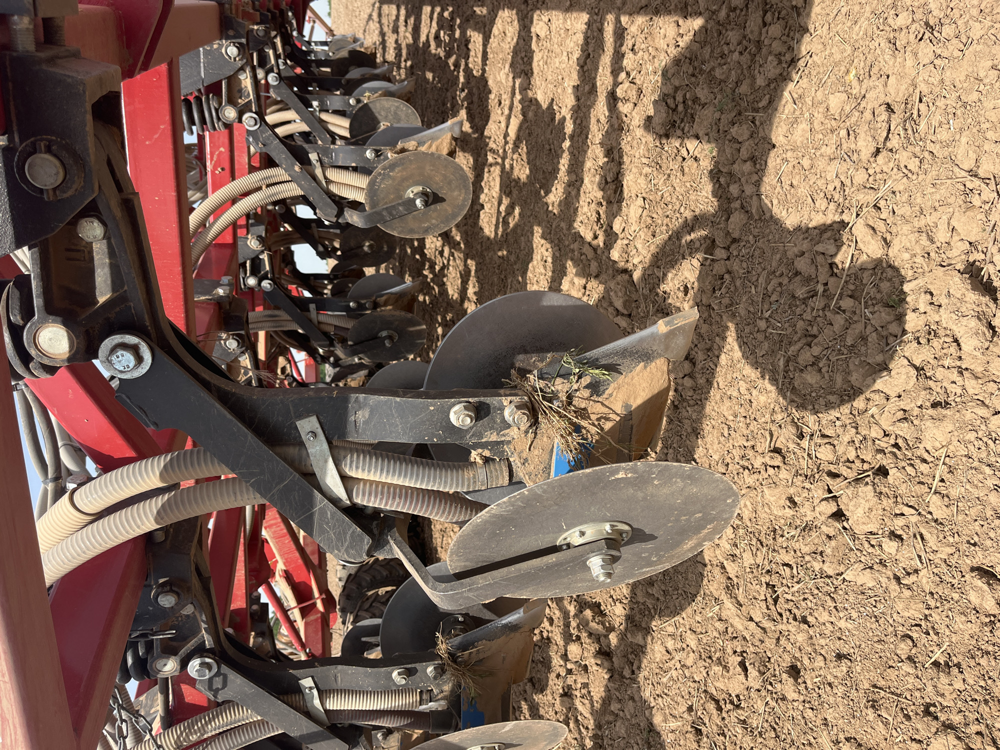 The image shows a close-up view of agricultural machinery, specifically a seeder or planter, positioned on tilled soil. The machinery features a series of discs or blades designed to create furrows in the soil for planting seeds. The components are a combination of red and black metal, with visible bolts, springs, and tubing. The soil is dry and cloddy, typical of freshly tilled land. The environment appears to be a rural setting, consistent with agricultural land. There are no people visible in the frame, but the shadow suggests a human presence. The location, city, and state are unknown.