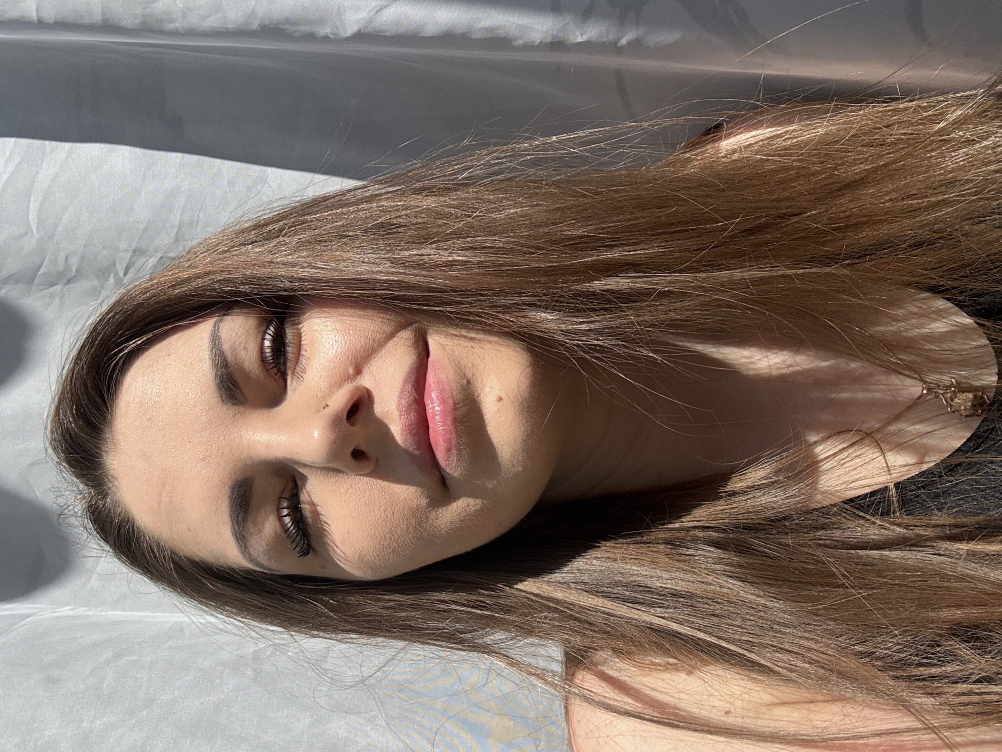 A young woman with long brown hair is lying on a white surface, possibly fabric. Her eyes are closed, and she has dark eyebrows and eyelashes. She has a nose ring. The lighting is strong, with clear shadows on her face and the surrounding surface. The scene appears to be indoors, possibly with natural light coming through a window. There is no evidence indicating the location is in Раздолье, Russia.