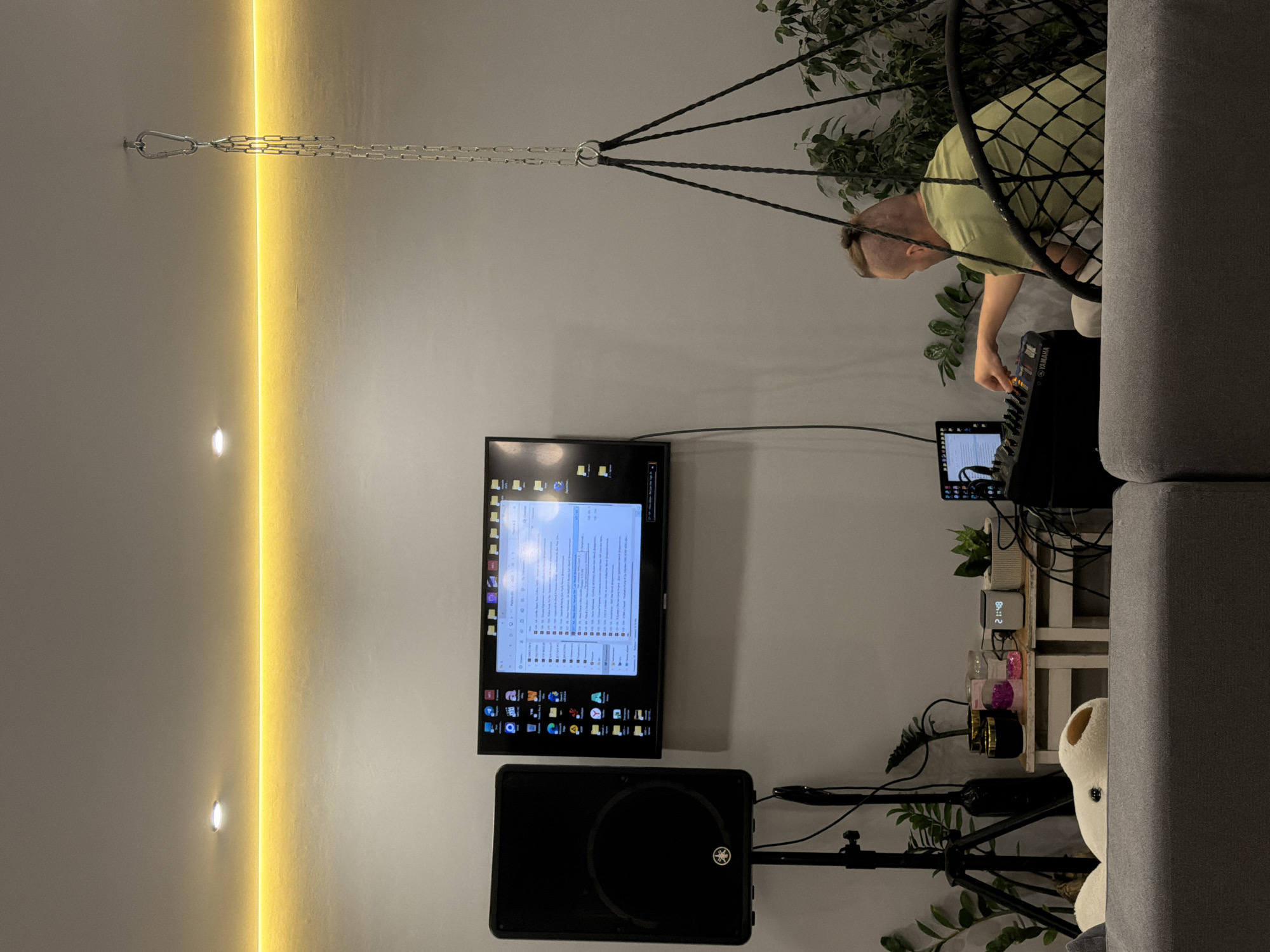 The image shows an indoor scene with a man adjusting a sound mixer while sitting in a hanging chair, likely in a private residence. He has a short, blond hairstyle with a shaved back. The chair is suspended from the ceiling by a chain. A black flat-screen TV is mounted on the wall, displaying a desktop interface with numerous application icons. Below the TV, there's a black Yamaha speaker on a stand. To the right of the speaker, a small table holds plants, a digital clock displaying "21:48," and various decorative items. A portion of a gray sofa is visible on the right side of the frame. An artificial plant is also near the right side of the image. The wall is painted a light color, and there's a vertical strip of warm, ambient lighting on the left side. The presence of the Yamaha speaker and mixing console suggest an audio setup, perhaps for music production or entertainment. No definitive evidence suggests the image was taken in Chaikovsky, Russia.