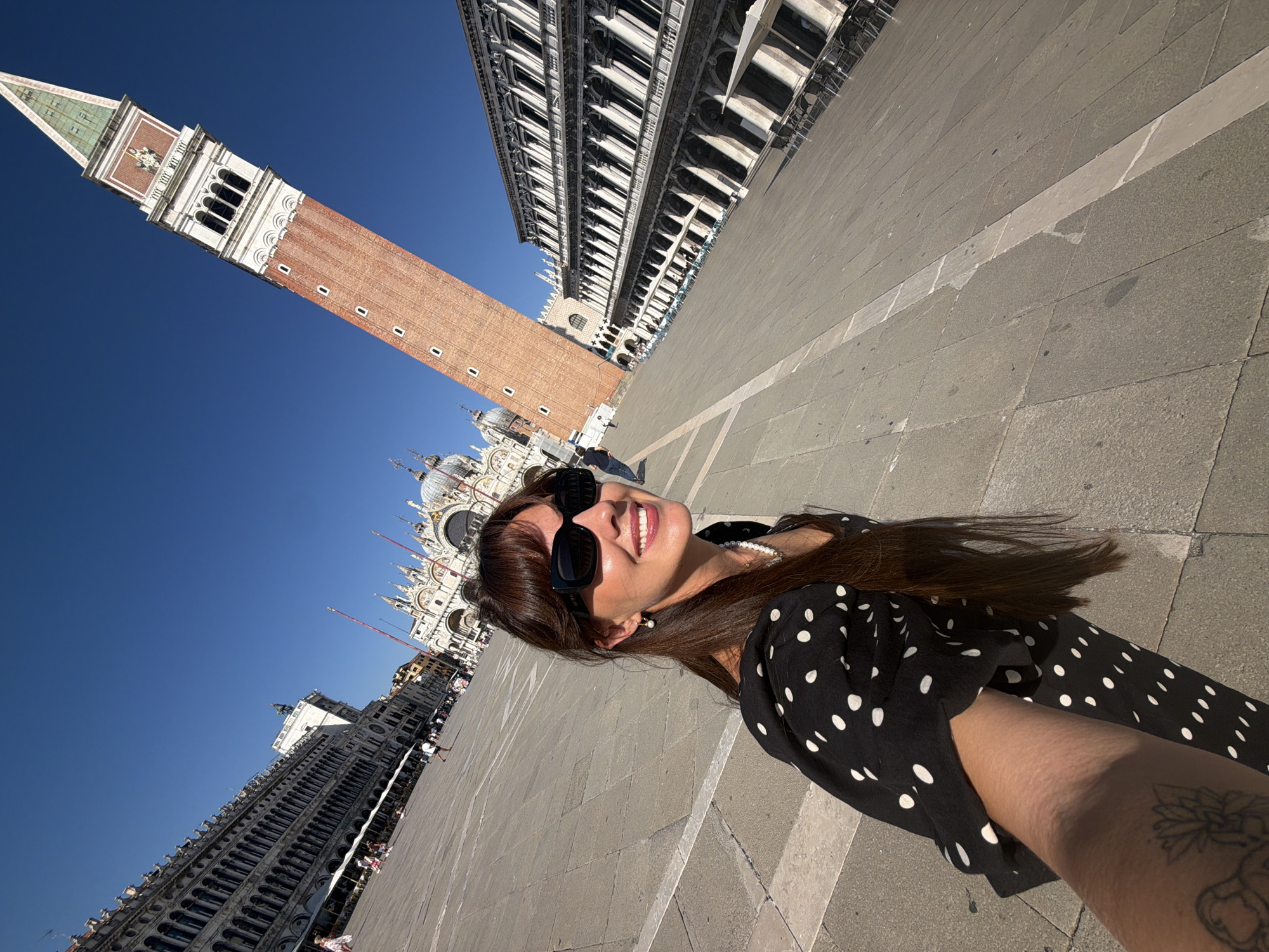 In Venice, Italy, a woman with long, dark hair and sunglasses takes a selfie in a vast, sunlit piazza. She is wearing a black dress with white polka dots and a pearl necklace. Behind her, the iconic Campanile di San Marco (St. Mark's Campanile) tower stands tall against a clear blue sky. The background also features the architectural grandeur of St. Mark's Basilica and the Procuratie buildings, with outdoor seating and umbrellas indicating a cafe or restaurant. The ground is paved with large, light-colored stones. People can be seen in the distance, walking and enjoying the square. The perspective is wide-angled, giving a sense of the open space.