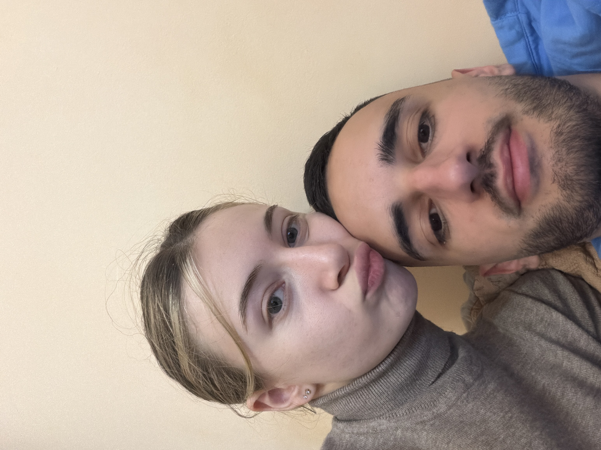 A selfie-style photograph captures a young woman and a young man posing closely together. The woman is positioned on the left side of the frame, her face angled slightly toward the camera with her lips pursed in a kissing gesture. She has fair skin and blond hair with darker roots. She wears a high-necked, neutral-toned sweater. The man is on the right side of the frame, his face close to the woman's. He has dark hair and a short, well-groomed beard. He appears to be wearing a blue shirt. The background is a plain, light yellow or beige wall. There are no identifiable landmarks or details to indicate a specific location, particularly not Plovdiv, Bulgaria.