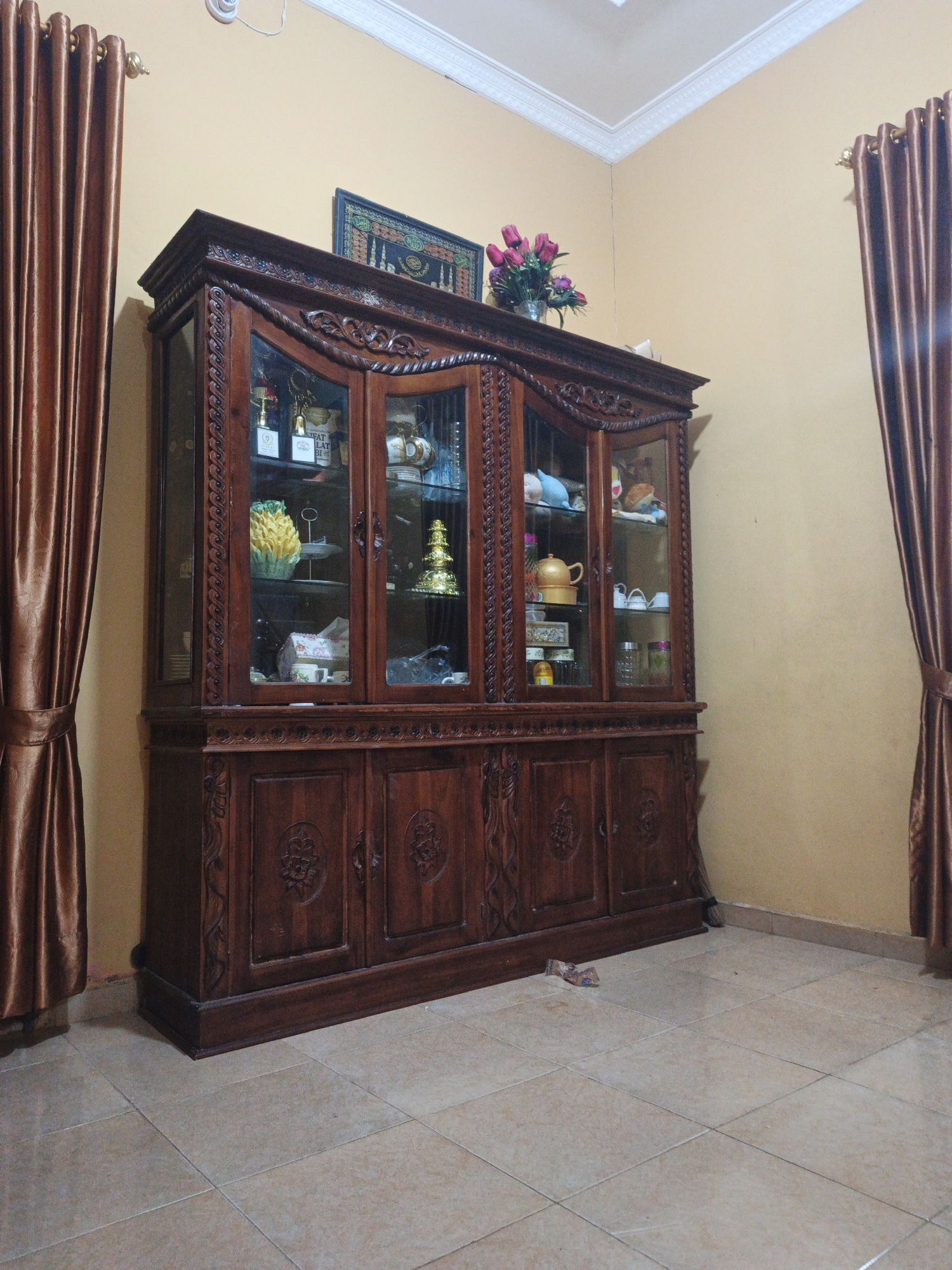 A dark wood, ornate china cabinet stands against a tan wall, flanked by brown satin curtains. The cabinet has glass doors revealing shelves filled with various decorative items and dishware. On top of the cabinet, a framed picture hangs on the wall and a vase of pink tulips is placed. The floor is tiled in a beige, square pattern. A piece of trash is visible on the floor near the cabinet. The scene appears to be an interior of a residential building in Pematang Duku, Indonesia.