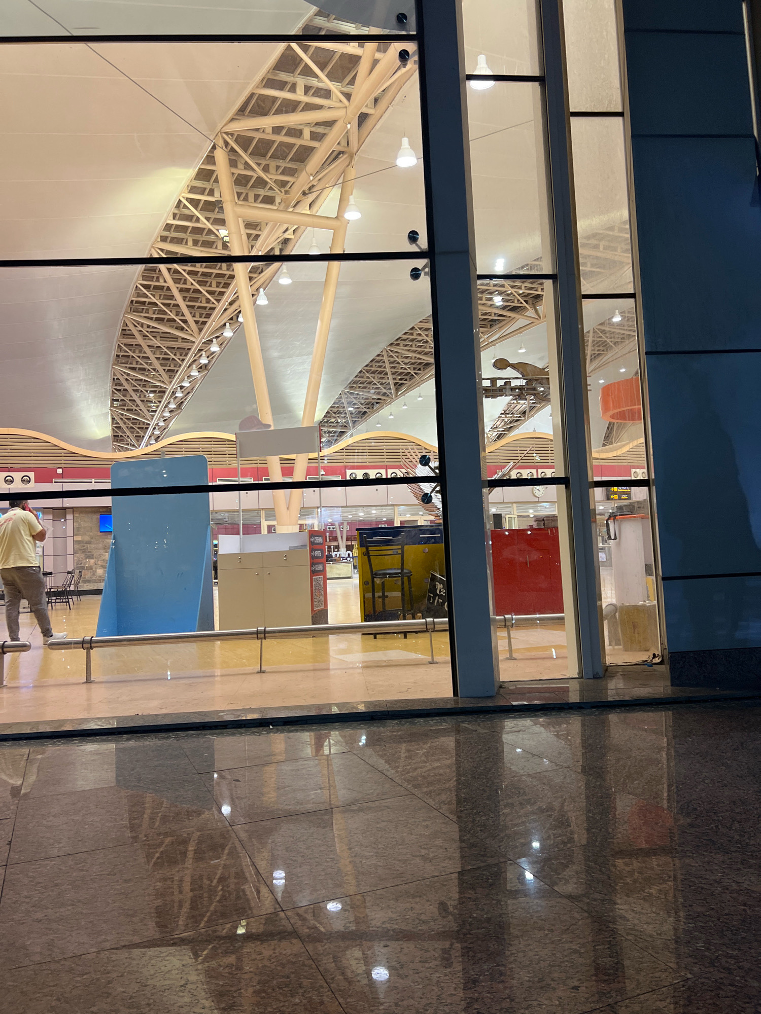The image shows the interior of an airport terminal, likely Sharm El Sheikh International Airport, Egypt, as seen through a large glass facade. The architecture features a high, curved ceiling supported by exposed beige metal beams and columns. The lighting consists of numerous white pendant lights along the ceiling. Inside the terminal, there are various elements visible: a person wearing a light-colored shirt and pants is walking away from the camera on the left. Several structural elements, including large blue and beige panels, a red partition, and a yellow desk with a chair, are positioned within the terminal. Traffic signs with directional arrows are also visible. The floor inside appears to be tiled and reflective, and the exterior ground also shows a wet, reflective surface, suggesting it may have recently rained or is being cleaned. The overall impression is that of a modern, well-lit airport at dusk or night.