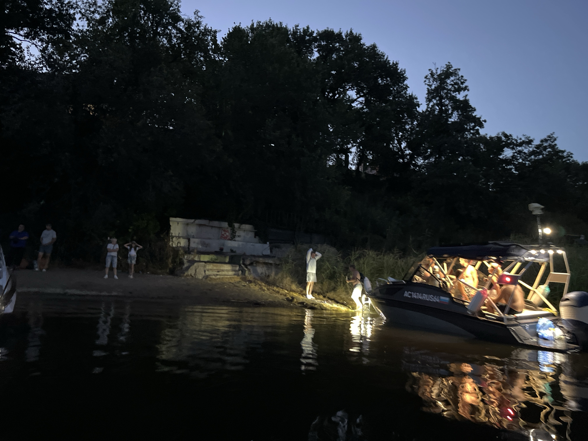 The image depicts a nighttime scene on the Volga River in Saratov, Russia. A group of people are gathered on the sandy shore, with several individuals standing near a boat. The boat, illuminated by interior lights, appears to be a powerboat with an outboard motor. The water reflects the lights of the boat and the surrounding environment. Trees line the background, silhouetted against the twilight sky. The overall impression is a relaxed social gathering by the river.
