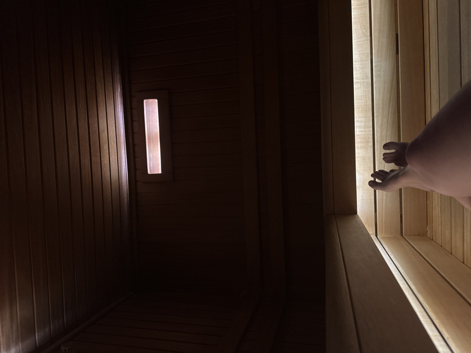 The scene is an interior view of a wooden sauna or similar facility located in Bartula, Montenegro. The room is constructed with dark-toned wooden slats on the walls and ceiling, and lighter natural wood slats forming multi-tiered benches. In the lower right quadrant, a pair of bare human feet and lower legs are visible, resting horizontally on a lower slatted bench. Subdued linear illumination emanates from beneath the edge of an upper bench, casting light onto the lower bench and the individual's feet. A horizontal rectangular opening, emitting a warm light, is present on the upper section of the back wall. The environment is dimly lit, with specific light sources providing localized illumination.