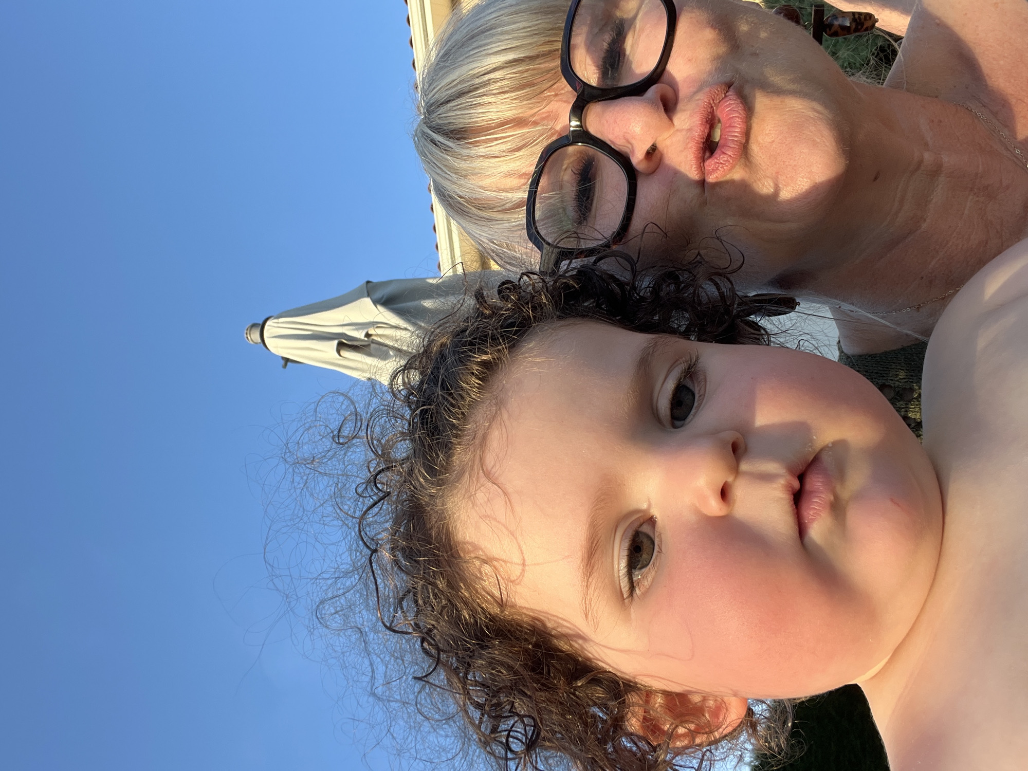 The image is a selfie taken outdoors with a clear blue sky in the background. In the foreground, two individuals are visible: a young child on the left and an adult on the right. The child's face is close to the camera, showing rosy cheeks and curly brown hair. The adult, on the right, is wearing glasses and making a kissing face. Behind them, part of a closed umbrella and a building's roofline are visible. The setting appears to be a residential area, possibly a backyard, given the umbrella and building. There is no specific indication in the image that this location is in the city of Saint-Galmier, France.