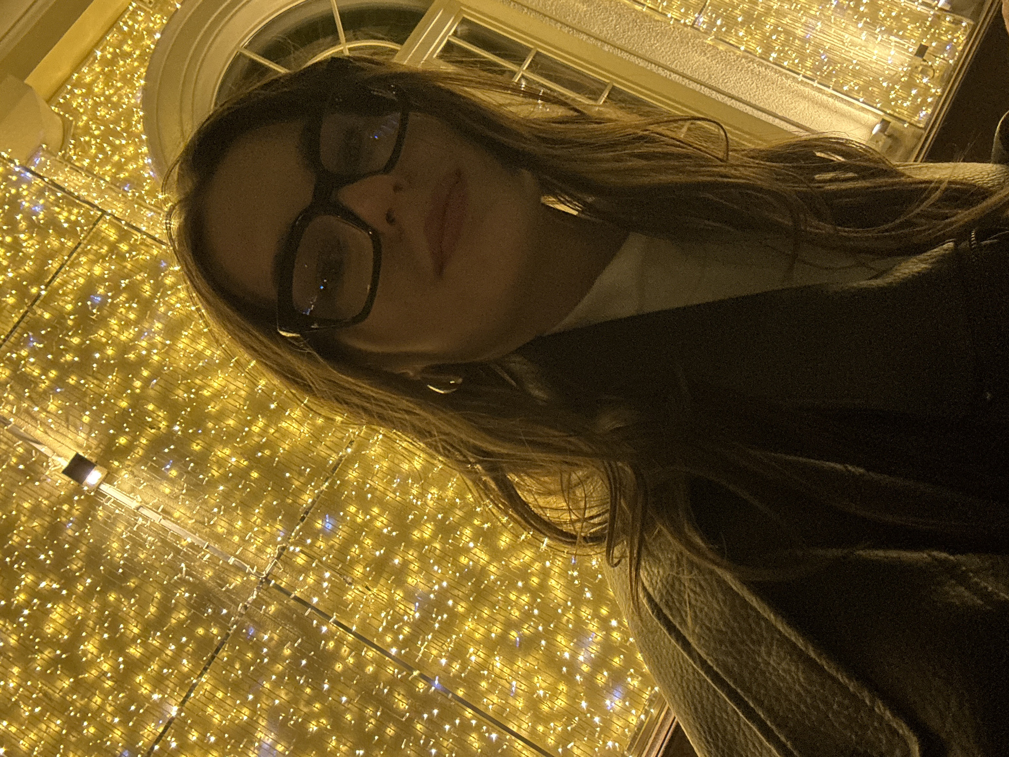 A woman with long brown hair and black-framed glasses is photographed at night in front of a decorative lighting arrangement. She is wearing a white turtleneck sweater and a dark-colored jacket. The background is filled with a grid-like pattern of small, warm-toned lights, likely part of an outdoor display. In the upper portion of the image, a rounded window frame can be seen above the lighting arrangement. The location is Moscow, Russia, indicated by the style of architecture and street decorations typical of the city during the holiday season.