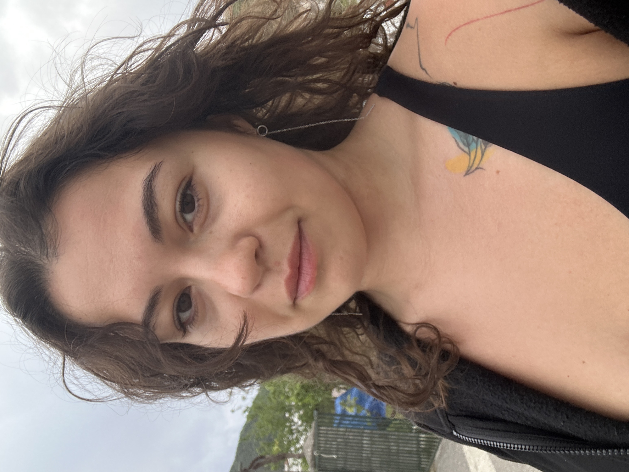 A selfie taken in Budva, Montenegro shows a woman with curly brown hair, wearing a black tank top and jacket. She has fair skin, brown eyes, and is wearing a delicate chain earring. Two tattoos are visible: one on her chest, and another on her upper back/shoulder. The background shows a cloudy sky, trees, and a blue object behind a fence, possibly indicating a construction site or a building under renovation. The photograph appears to be taken outdoors during the day.