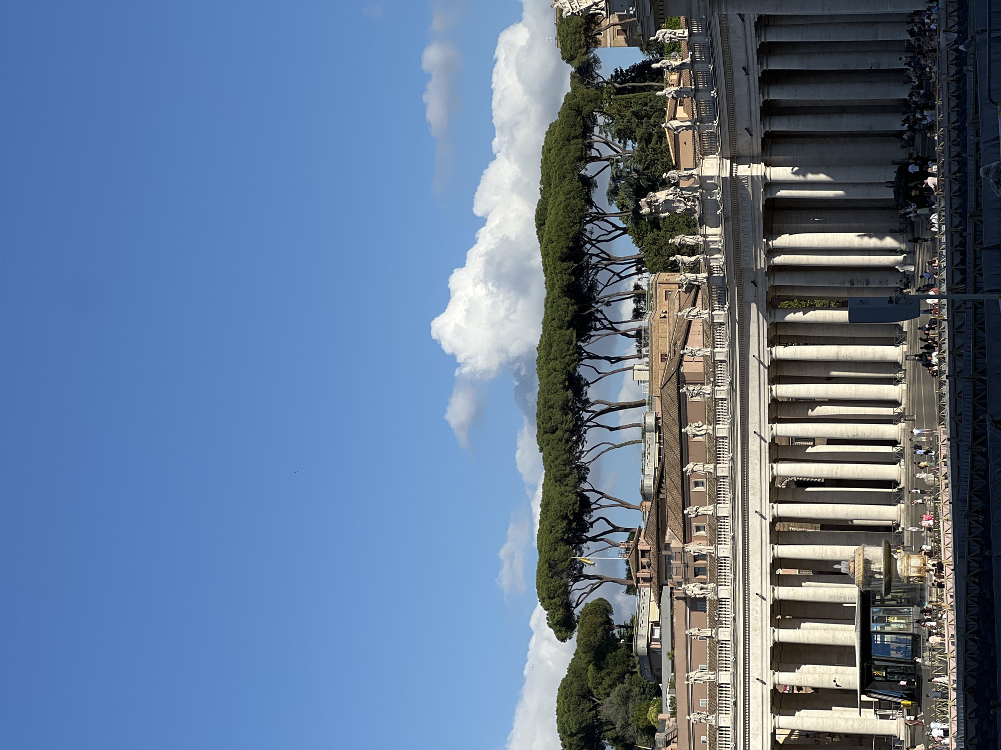 The image depicts St. Peter's Square in Città del Vaticano, Italy, under a clear blue sky with scattered cumulus clouds. In the midground, a large semicircular Doric colonnade extends across the frame, composed of multiple rows of light-colored stone columns. An entablature above the columns features an inscription that includes "ALEXANDER VII PONT. MAX." Numerous statues of saints are positioned atop the colonnade.
In the foreground of the colonnade, a modern, rectangular glass structure with a dark roof is visible on the left, and a multi-tiered stone fountain actively spraying water is situated to its right. A dark, box-like object stands in the lower-left foreground near the colonnade. The square in front of the colonnade is populated by numerous individuals, some walking, others standing or gathered behind a low, dark barrier in the immediate foreground. Long, distinct shadows are cast by the columns, indicating a low sun angle.
The background reveals a line of distinctive umbrella pine trees with tall, slender trunks and flattened green canopies, appearing to crest a ridge or hill. Behind and below these trees, various multi-story buildings with light-colored facades and reddish-brown tiled roofs are visible. A yellow flag, likely that of Vatican City, flies from a flagpole positioned amidst the pine trees. The upper portion of the frame is dominated by the blue sky and white clouds, with a tiny, dark object (possibly a bird or distant aircraft) visible in the upper-right.